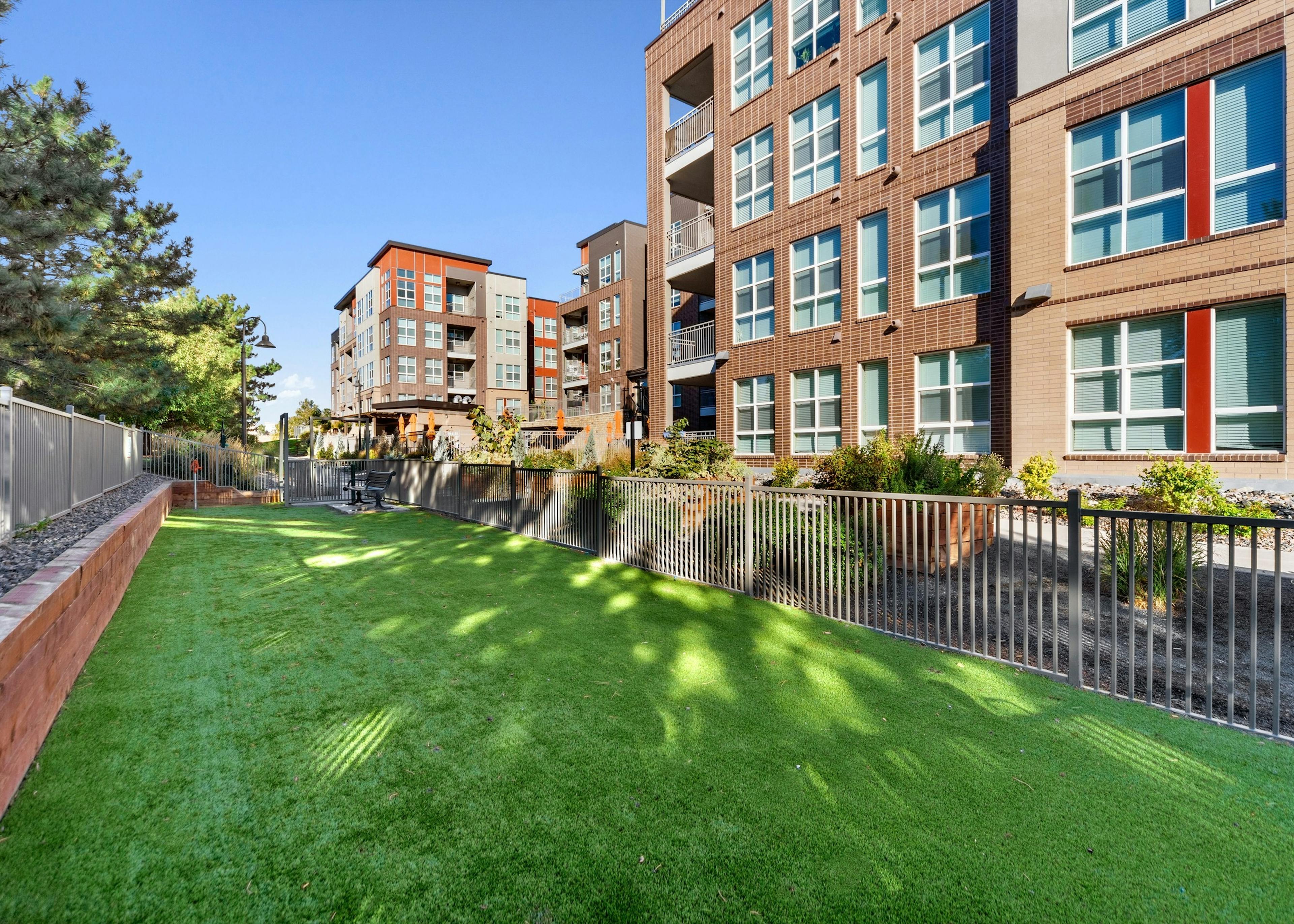 A view at the paw park at AMLI Dry Creek apartments with green grass and bench at far with  surrounding fence and brick building in rear