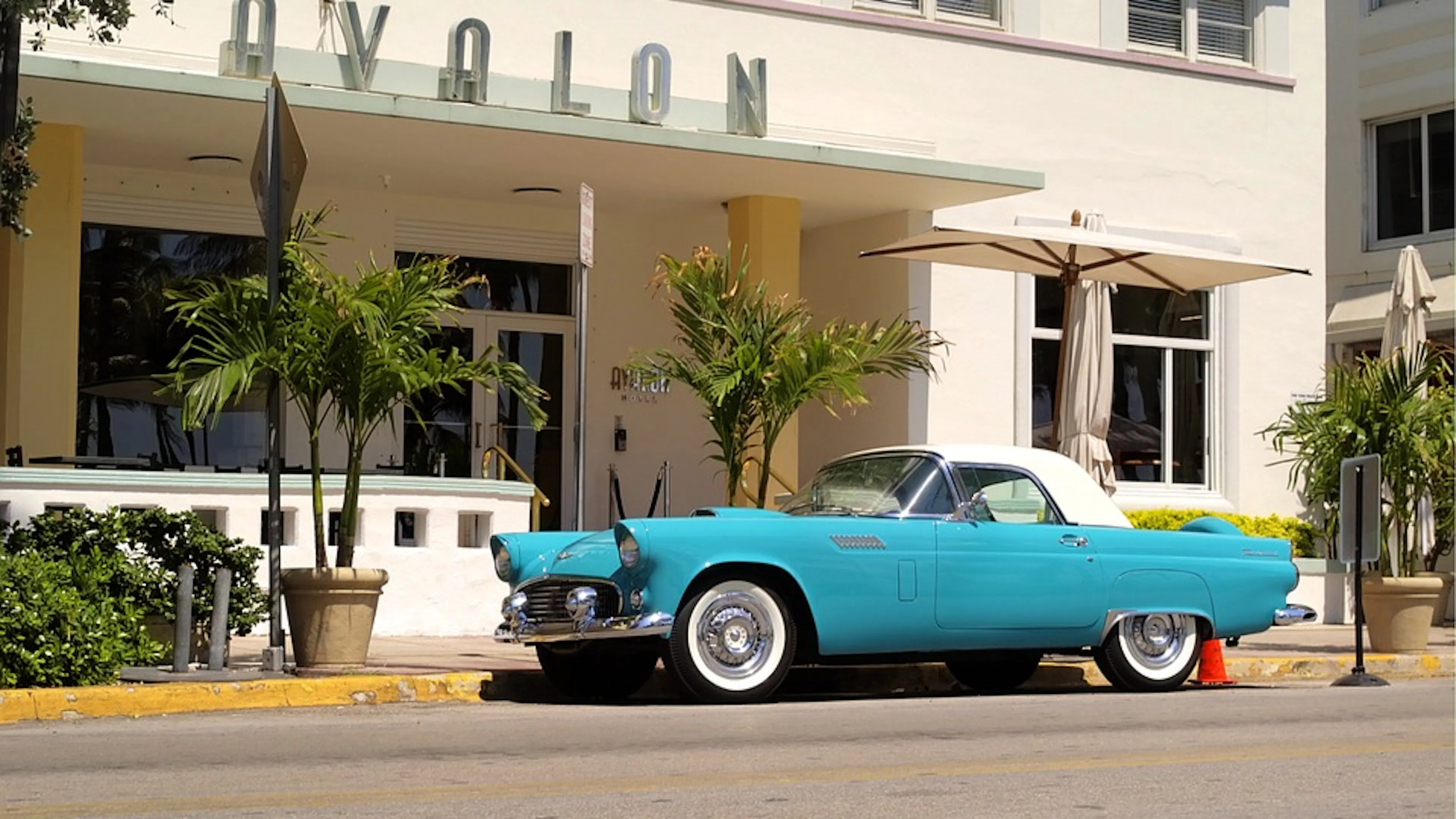 A blue vintage car parked outside a vintage hotel surrounded by palm trees and plenty of sunshine