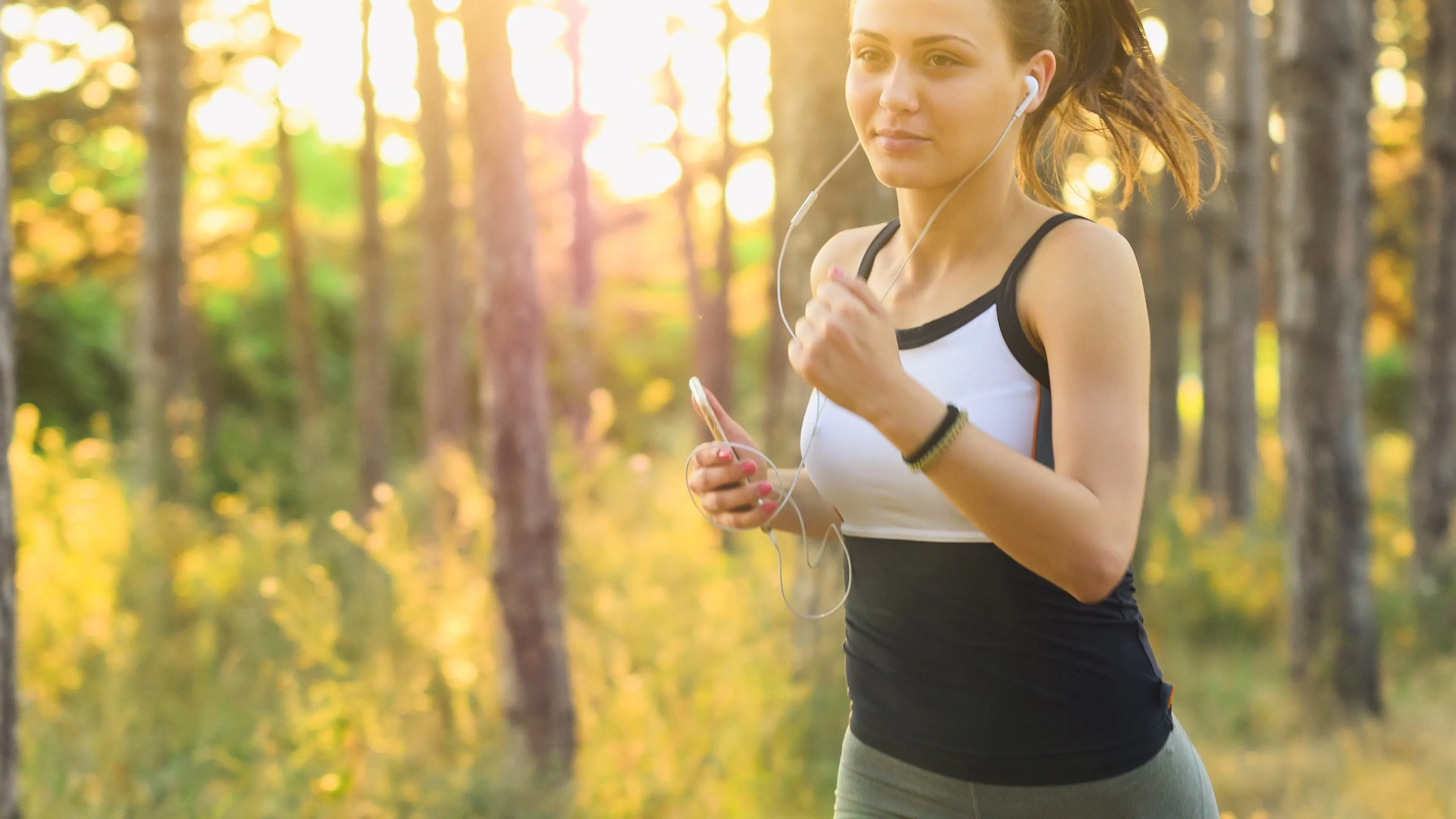 woman with white and black top and earbuds running by tress and yellow foliage