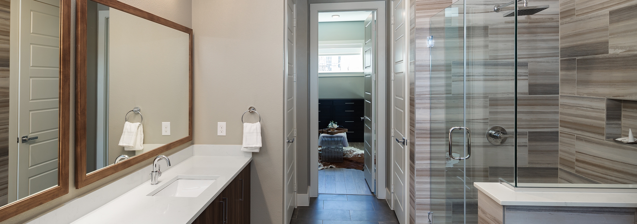 Interior of AMLI South Shore bathroom with glass shower with grey and tan wall tiles and a vanity sink with peek into bedroom