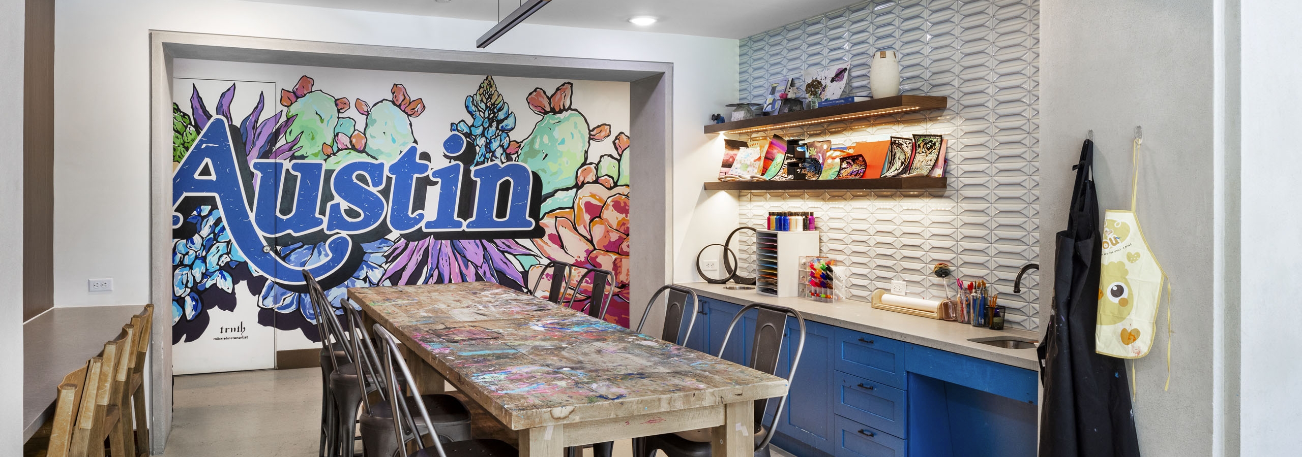 AMLI Lakeline maker space with a colorful Austin mural and a long wooden table flanked by metal chairs and blue cabinetry