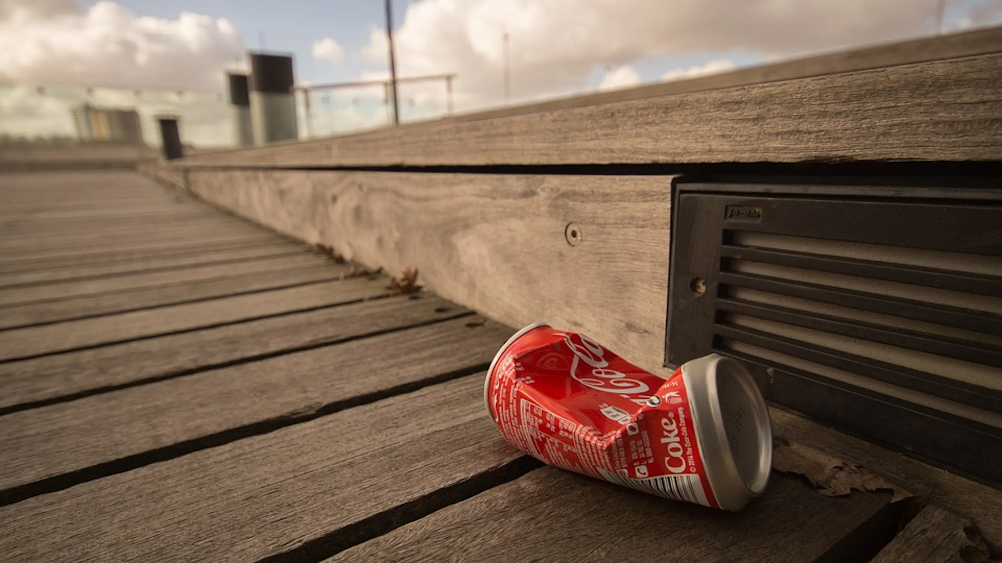 A close up view of a crushed Coca Cola can resting on  wooden boardwalk in what appears to be a city 