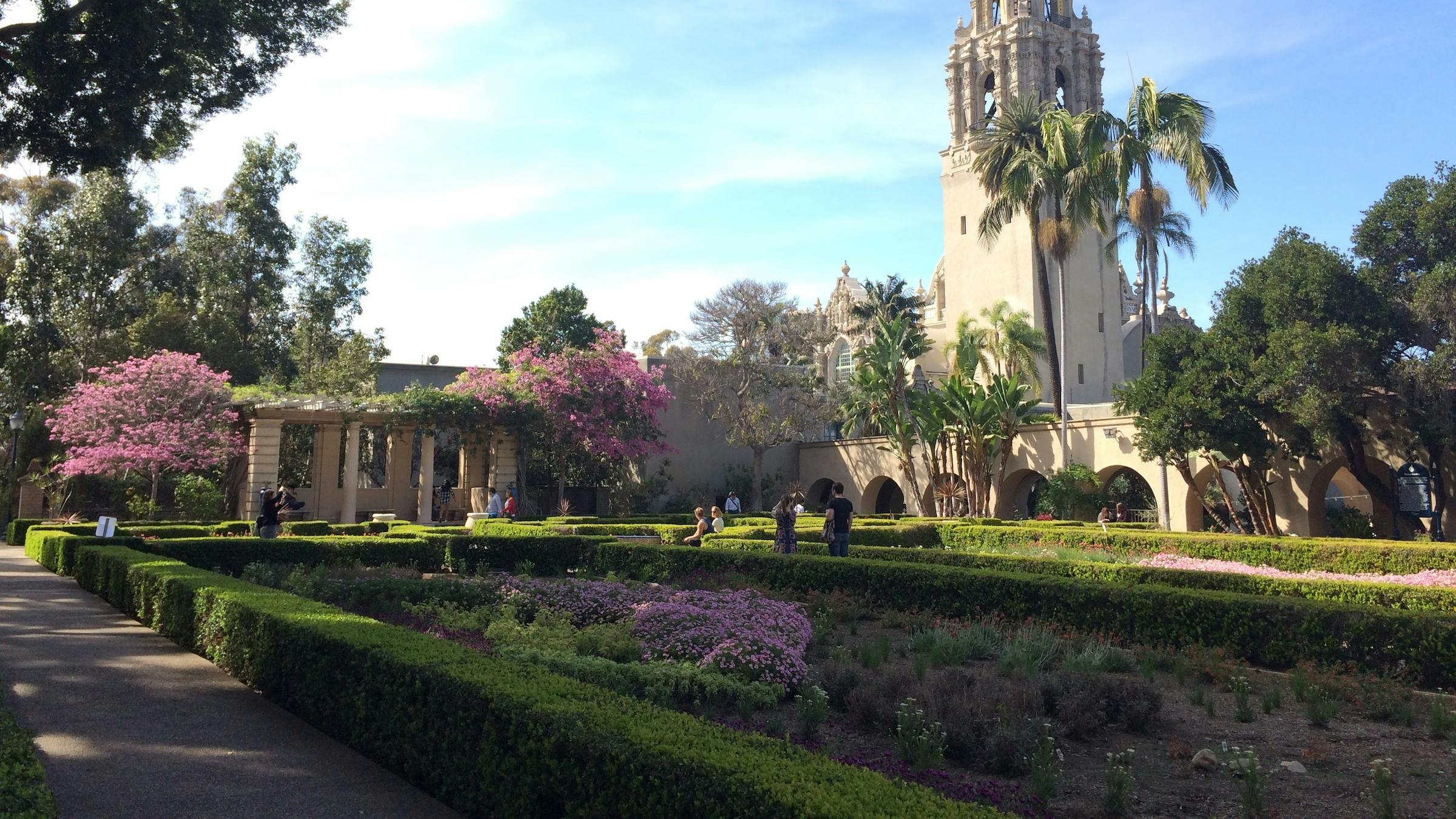 Daytime view of Balboa Park with green hedges, trees, walking path, and the California Tower visible.