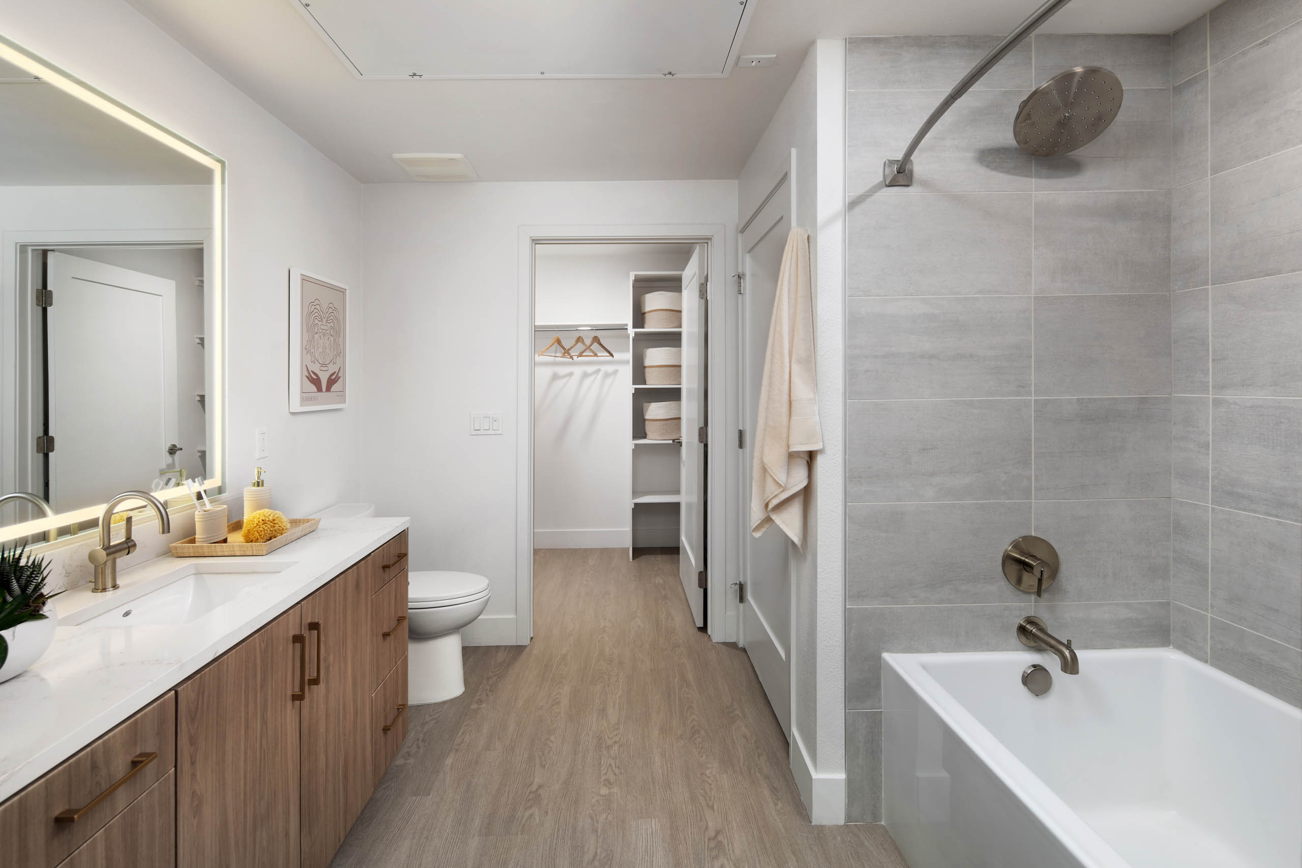 Modern bathroom with wood vanity, lit mirror, white countertop, toilet, gray tiled tub, large showerhead, and towel.