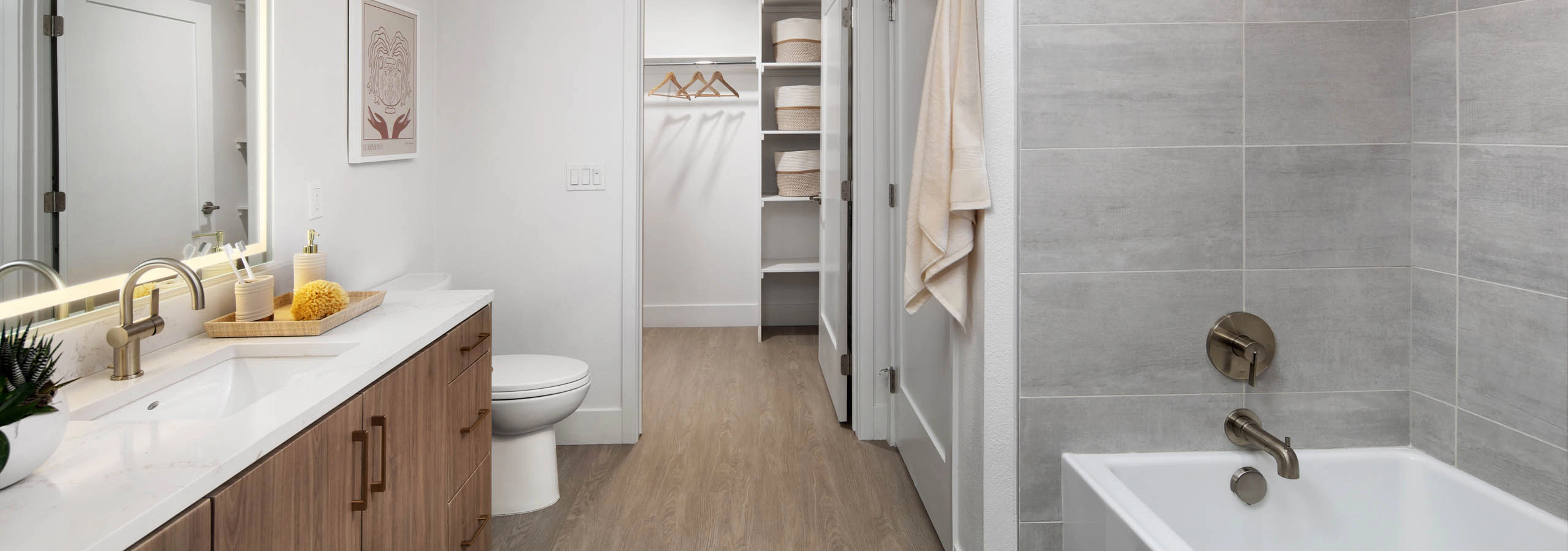 Modern bathroom with wood vanity, lit mirror, white countertop, toilet, gray tiled tub, large showerhead, and towel.