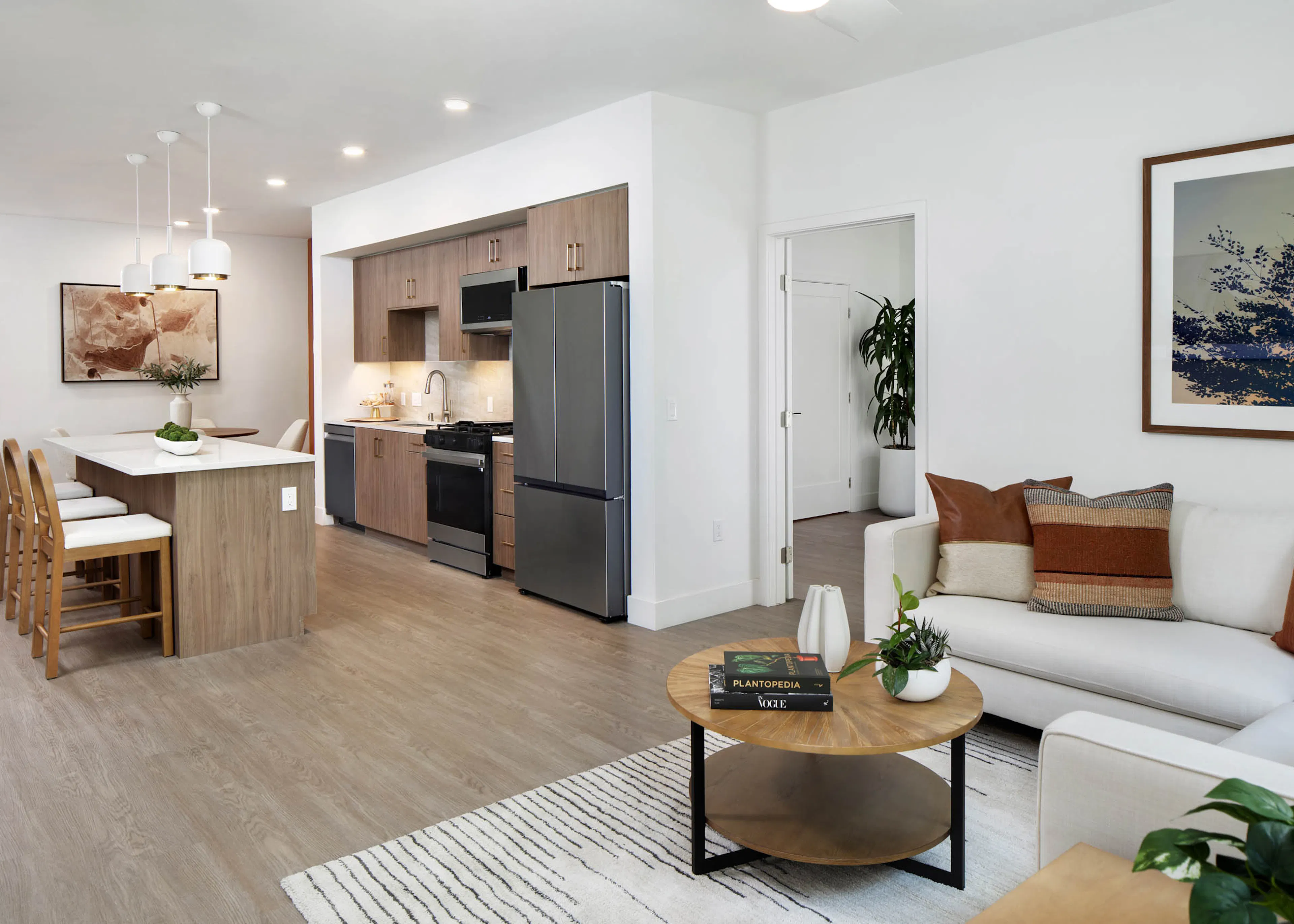 Open living room and kitchen with white sofa, round coffee table, wood cabinets, island seating, pendant lights, framed art, and plants.
