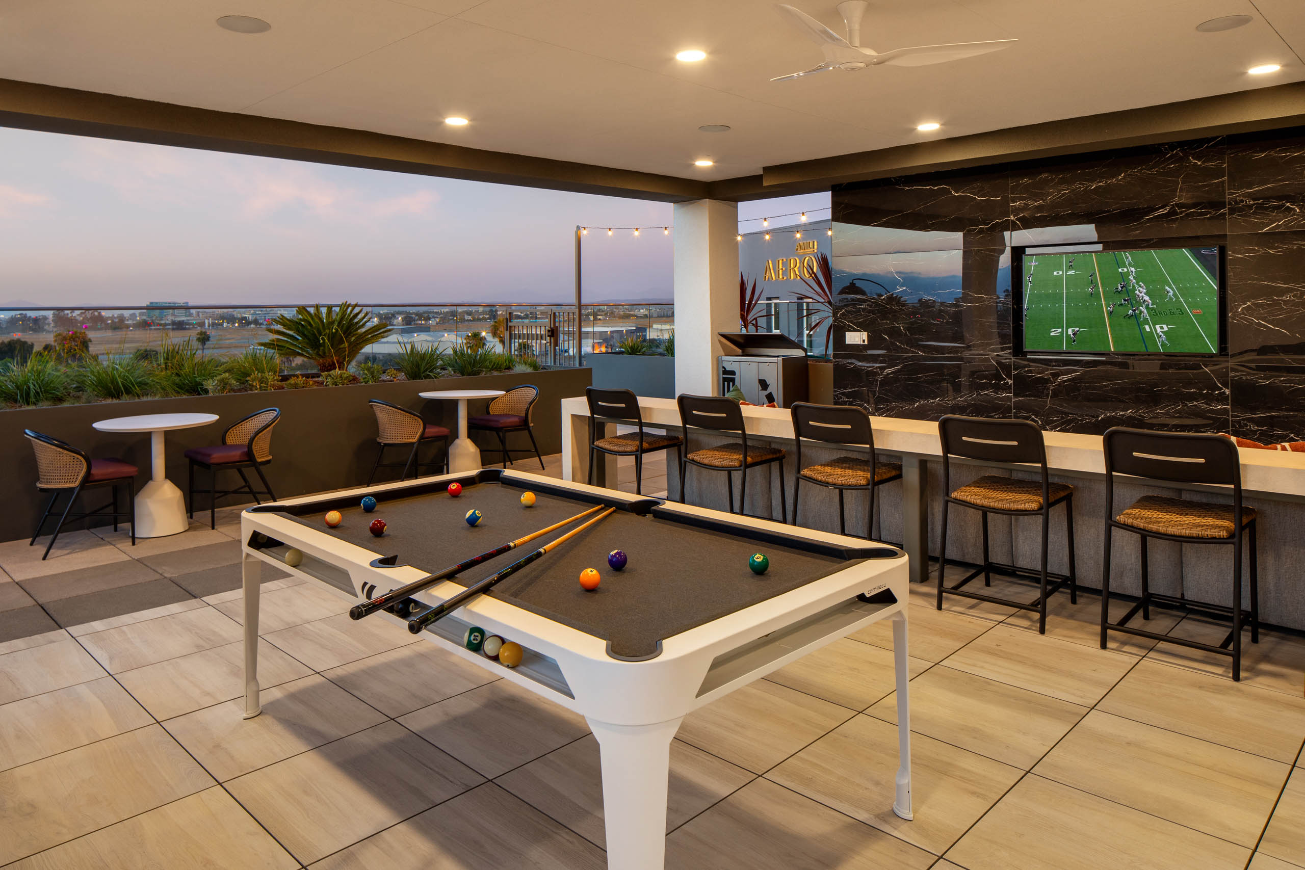Outdoor lounge and entertainment area with pool table, bar seating, wall-mounted TV, small tables, rooftop views, and open-air covered patio.