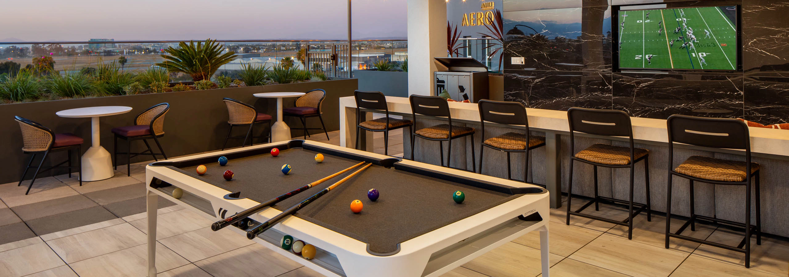 Outdoor lounge and entertainment area with pool table, bar seating, wall-mounted TV, small tables, rooftop views, and open-air covered patio.