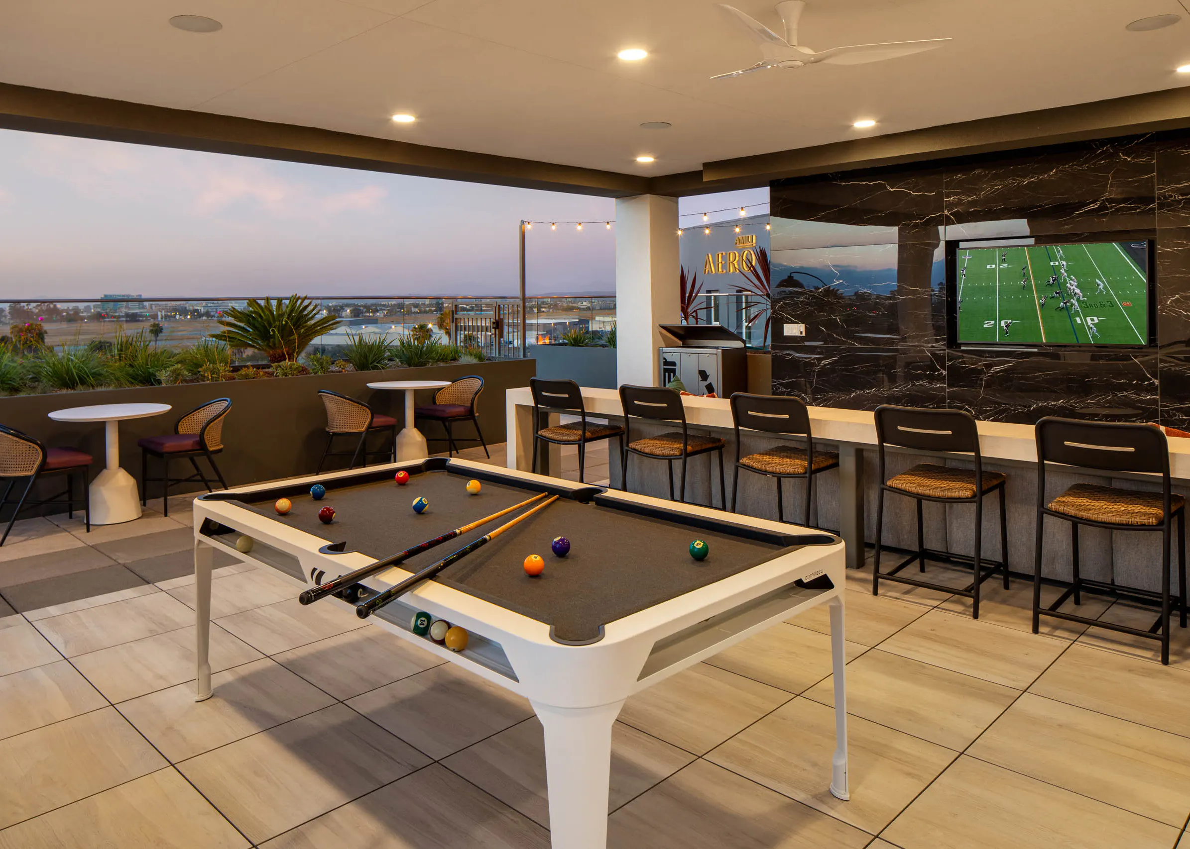 Outdoor lounge and entertainment area with pool table, bar seating, wall-mounted TV, small tables, rooftop views, and open-air covered patio.