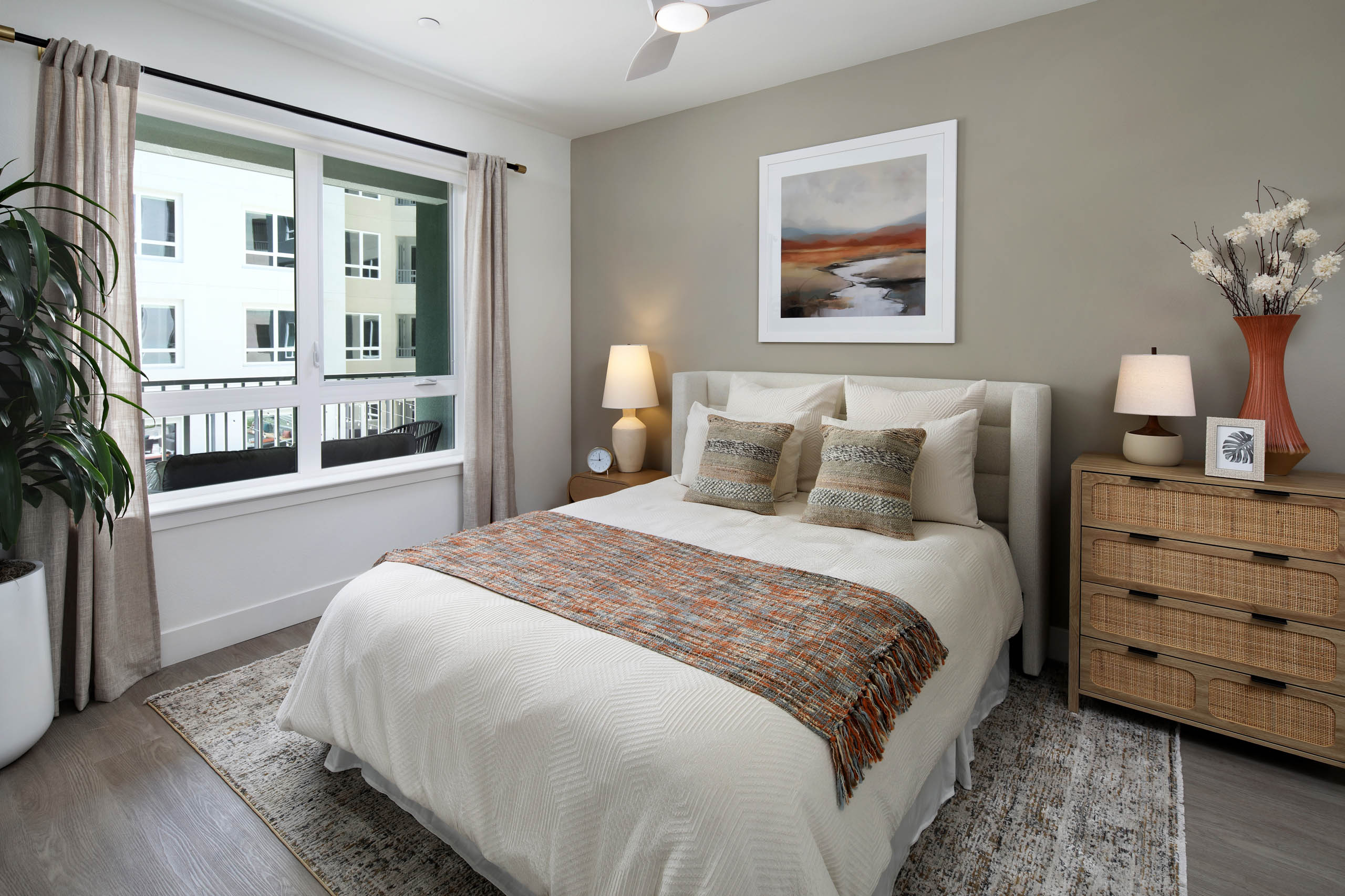 Bedroom with white bedding, upholstered headboard, large window with curtains, ceiling fan, plant, landscape art, lamps, and woven-front dresser.