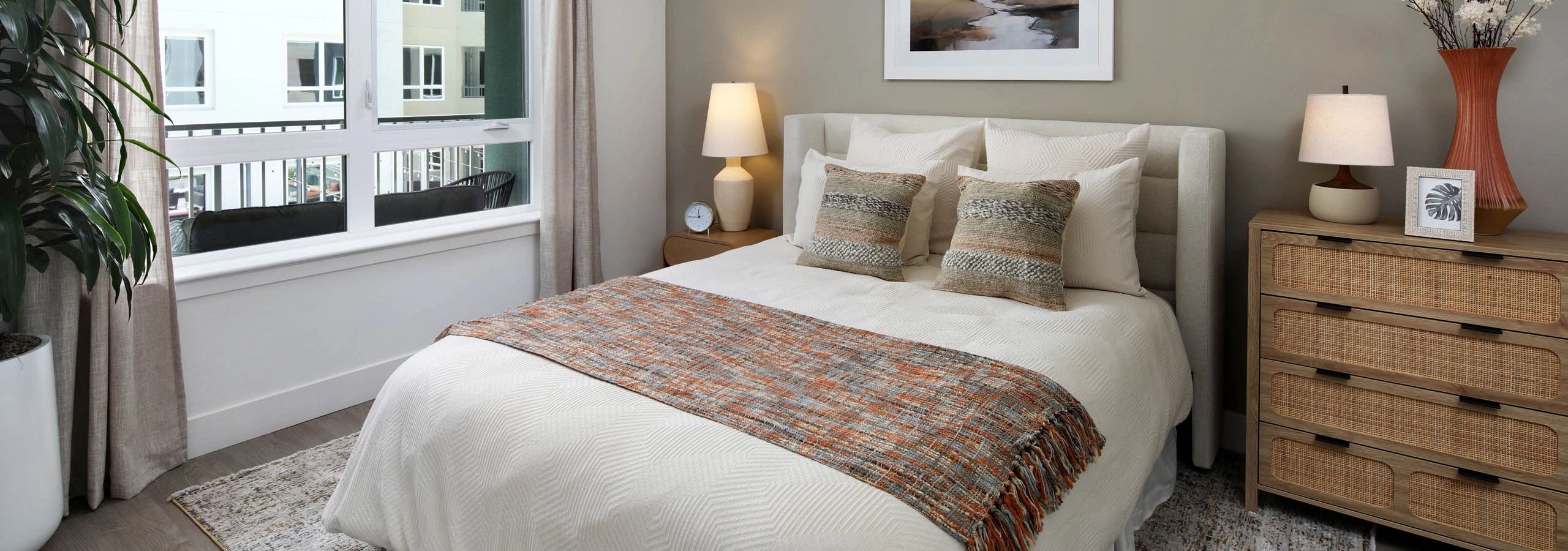 Bedroom with white bedding, upholstered headboard, large window with curtains, ceiling fan, plant, landscape art, lamps, and woven-front dresser.