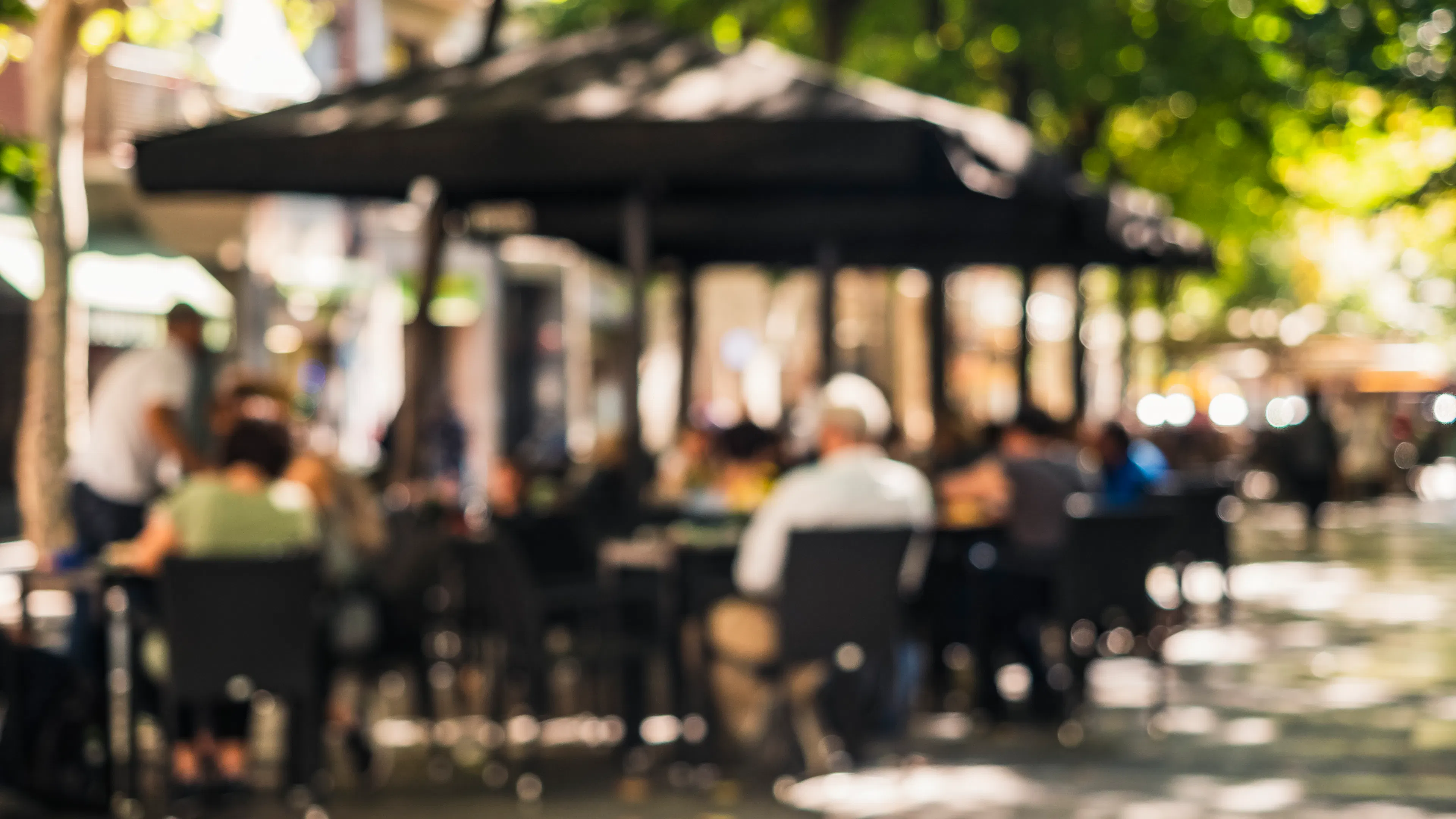 Blurred outdoor patio dining area with people enjoying lunch under shade umbrellas