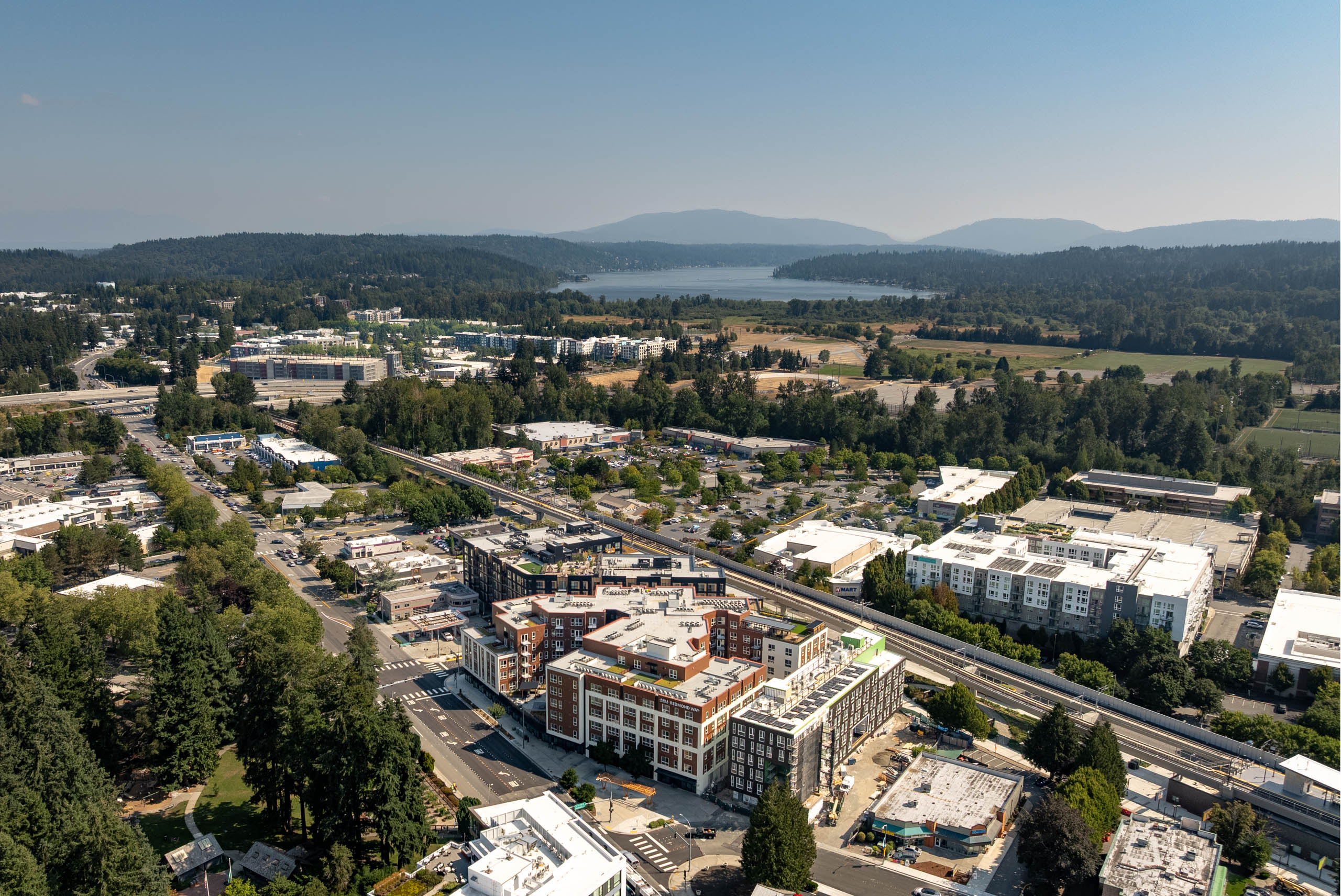Wide aerial view of AMLI Redmond Way and downtown Redmond, with the light rail line, surrounding neighborhoods, forested hills, and a lake visible in the distance.