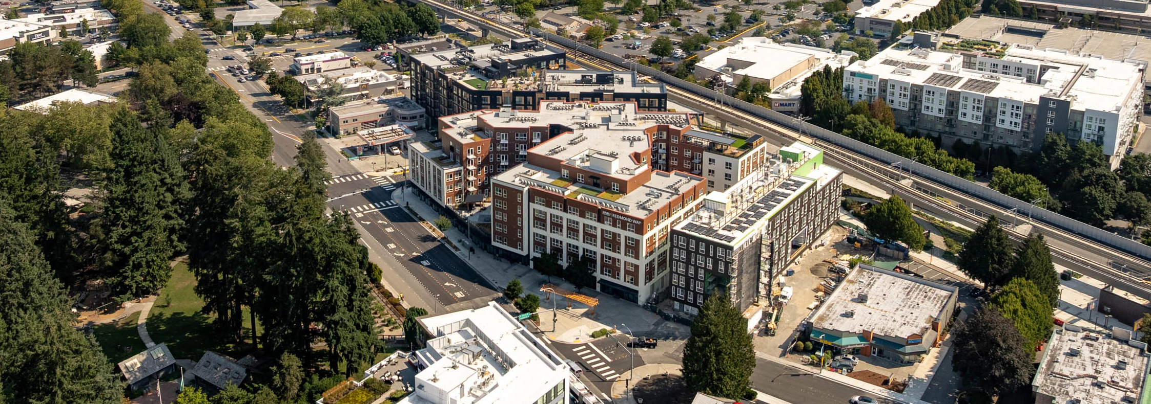 Wide aerial view of AMLI Redmond Way and downtown Redmond, with the light rail line, surrounding neighborhoods, forested hills, and a lake visible in the distance.