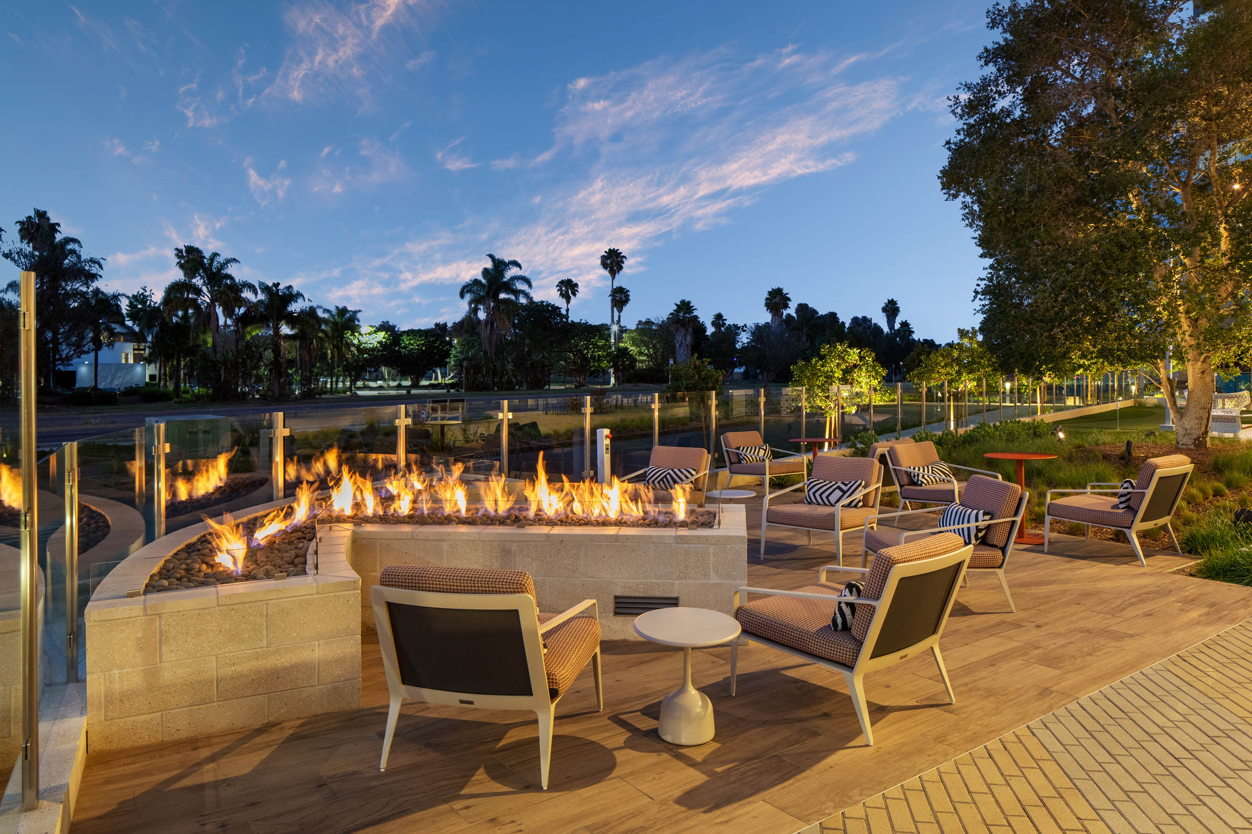 Outdoor fire lounge with cushioned seating, fire feature, side tables, landscaped grounds, glass fencing, palm trees, and evening sky views.
