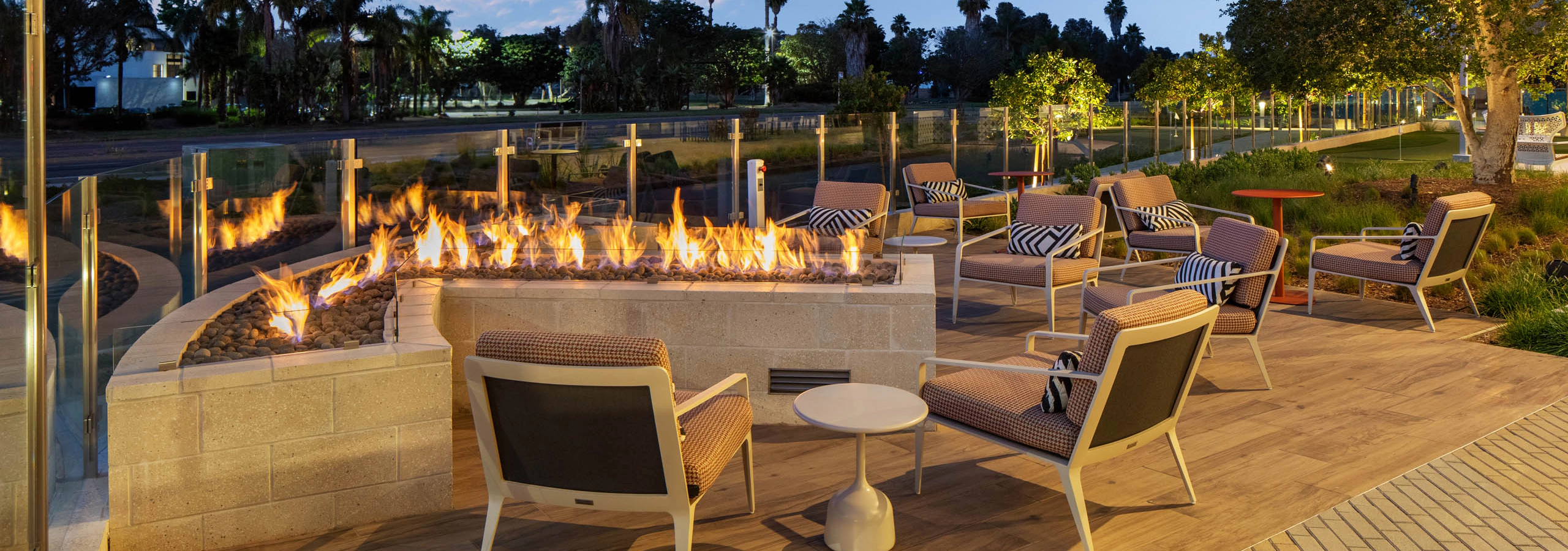Outdoor fire lounge with cushioned seating, fire feature, side tables, landscaped grounds, glass fencing, palm trees, and evening sky views.