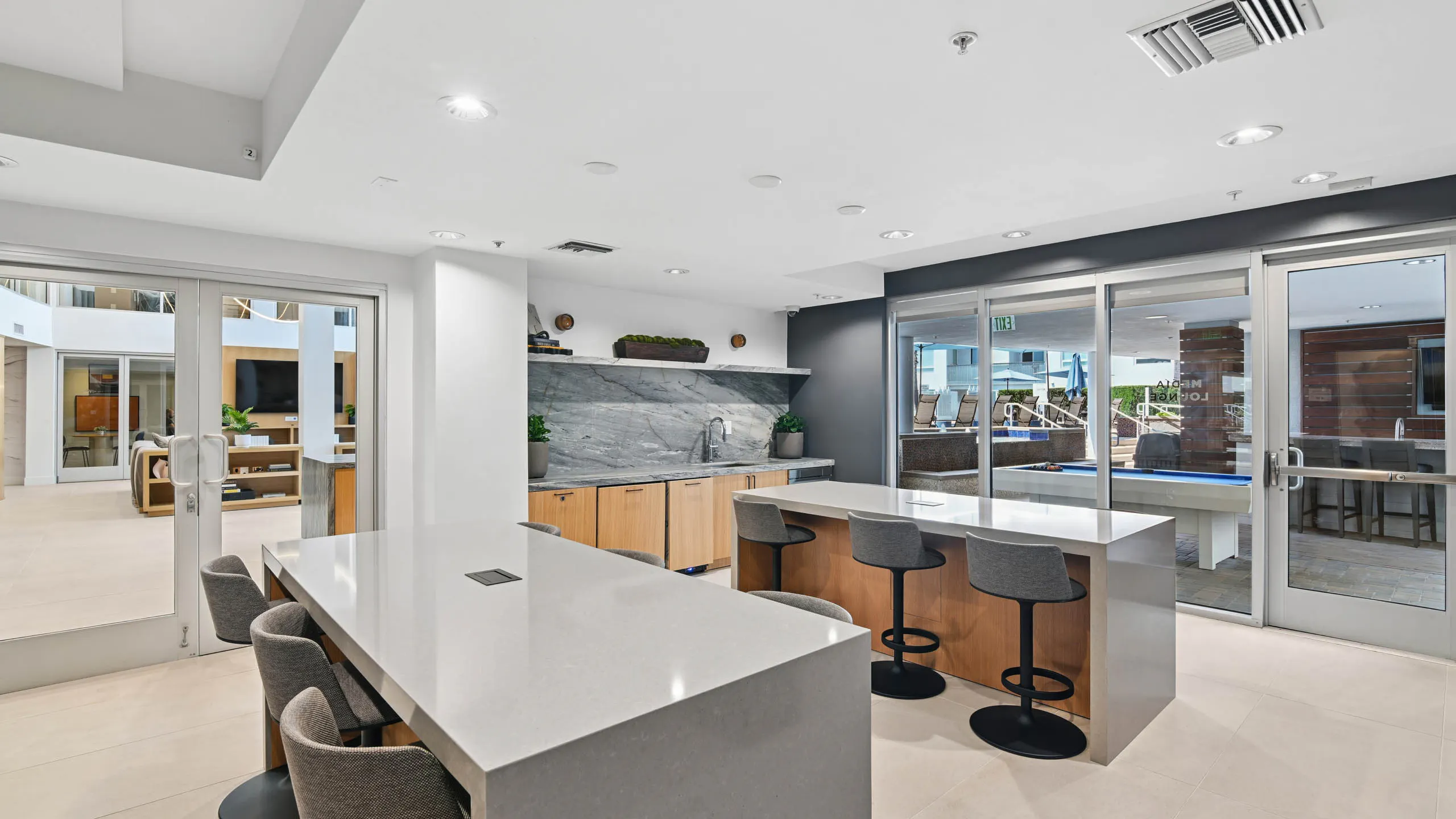 Media lounge at AMLI Lex on Orange with two counter-height tables with gray stools, a counter with sink, stone backsplash, and wood cabinets, and glass doors to an outdoor area with a pool table.