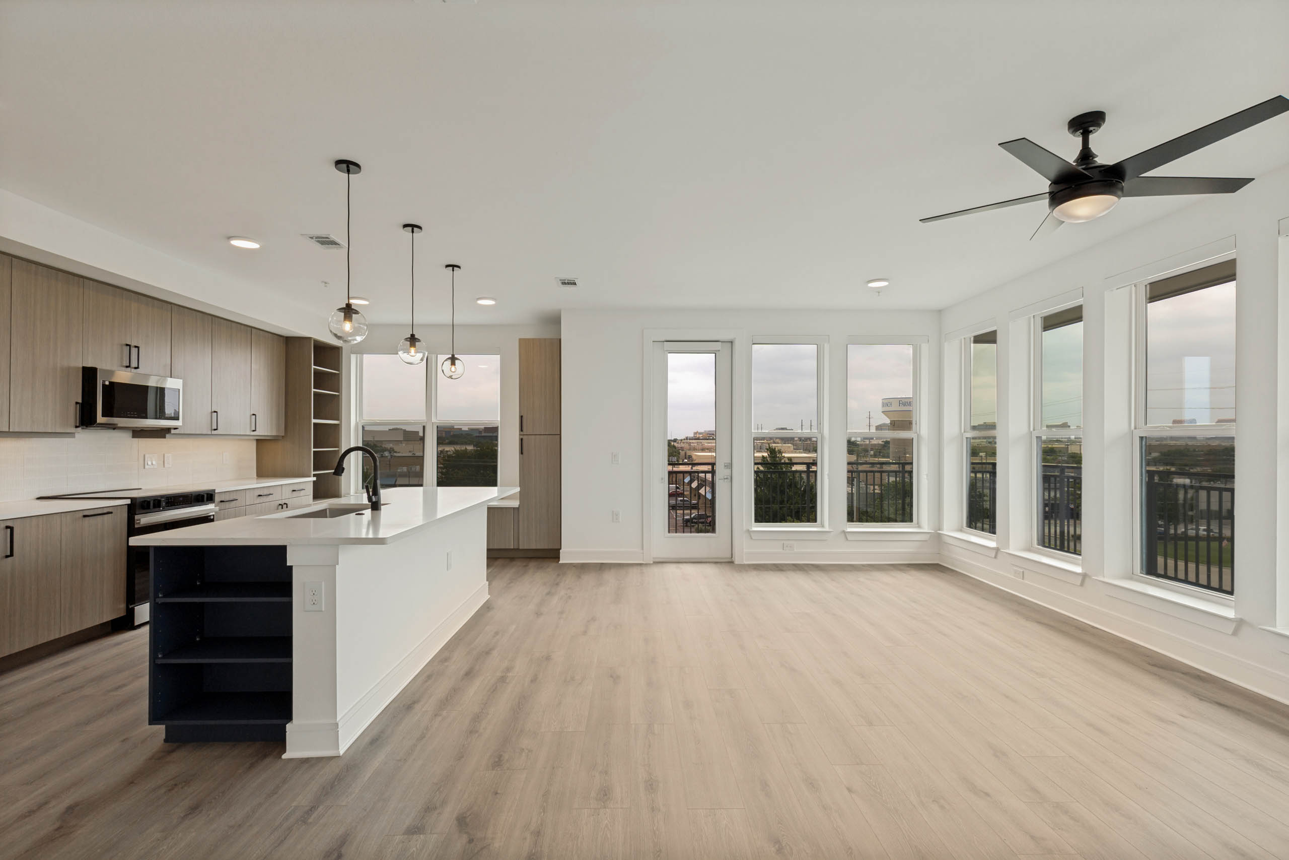 Spacious open-concept living area with kitchen island, wood-style flooring, pendant lighting, balcony access, and large windows.