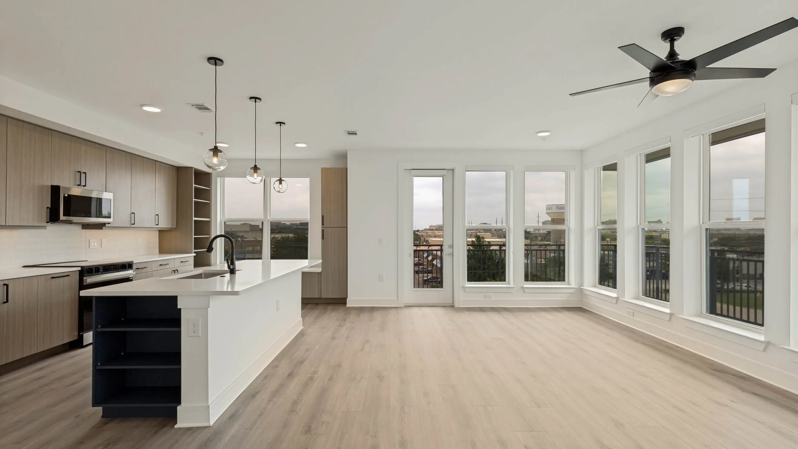 Spacious open-concept living area with kitchen island, wood-style flooring, pendant lighting, balcony access, and large windows.