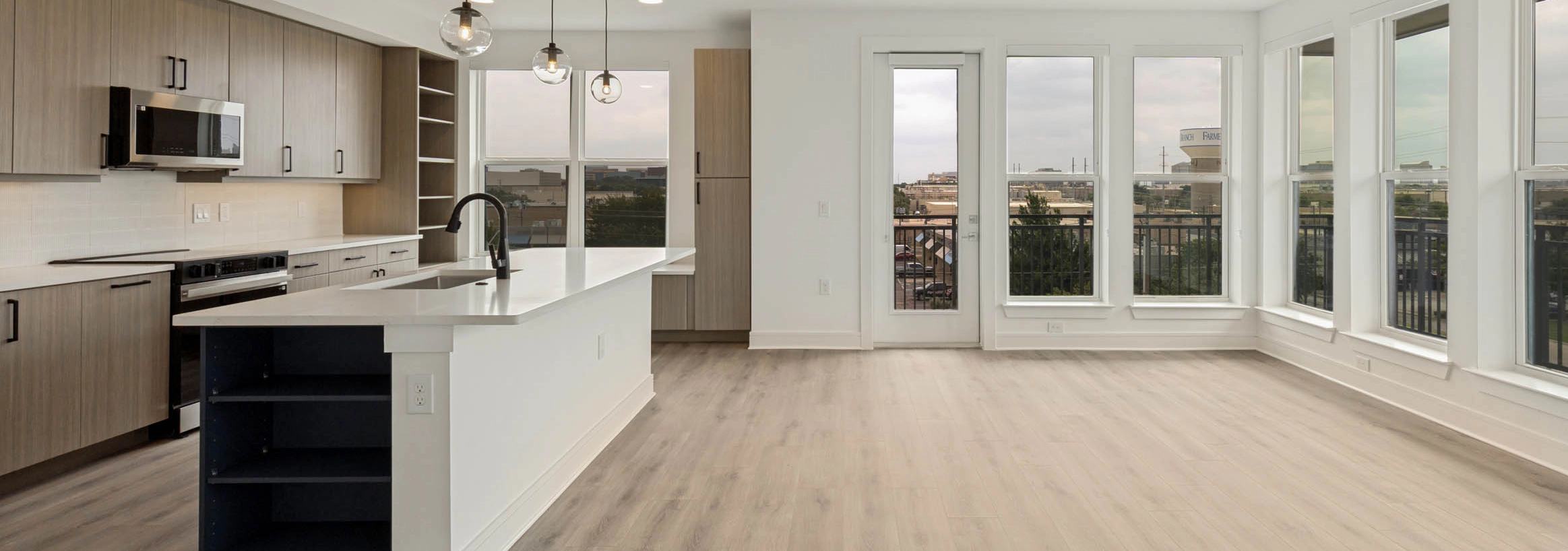 Spacious open-concept living area with kitchen island, wood-style flooring, pendant lighting, balcony access, and large windows.