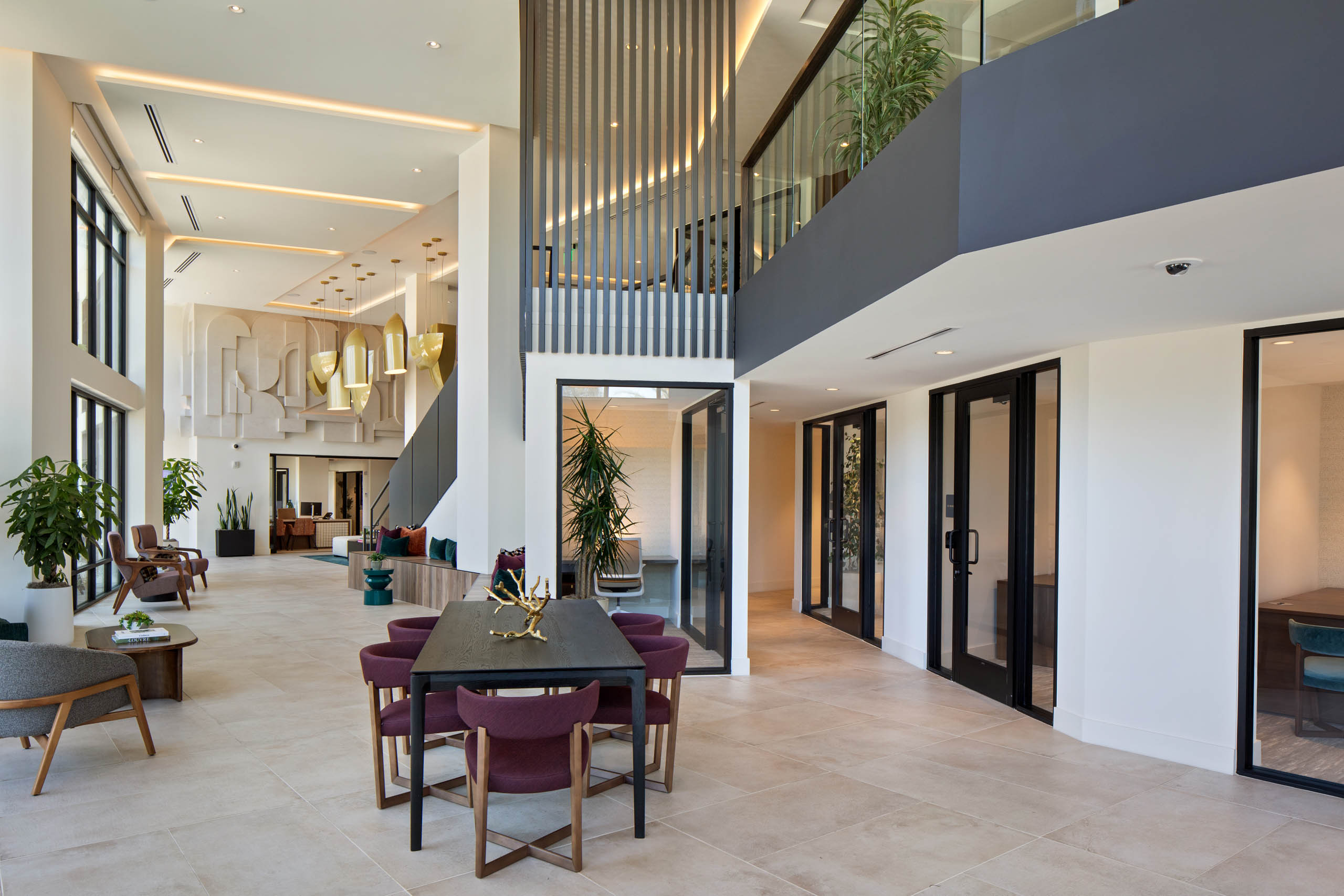 Modern two-story lounge with seating, conference rooms, large windows, pendant lighting, decorative wall art, plants, and a communal table.