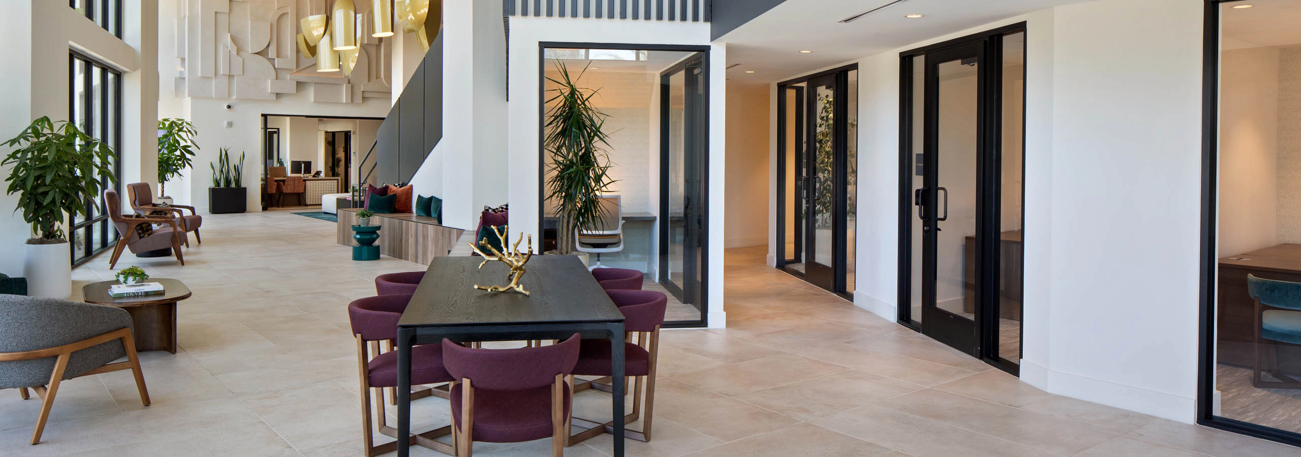 Modern two-story lounge with seating, conference rooms, large windows, pendant lighting, decorative wall art, plants, and a communal table.