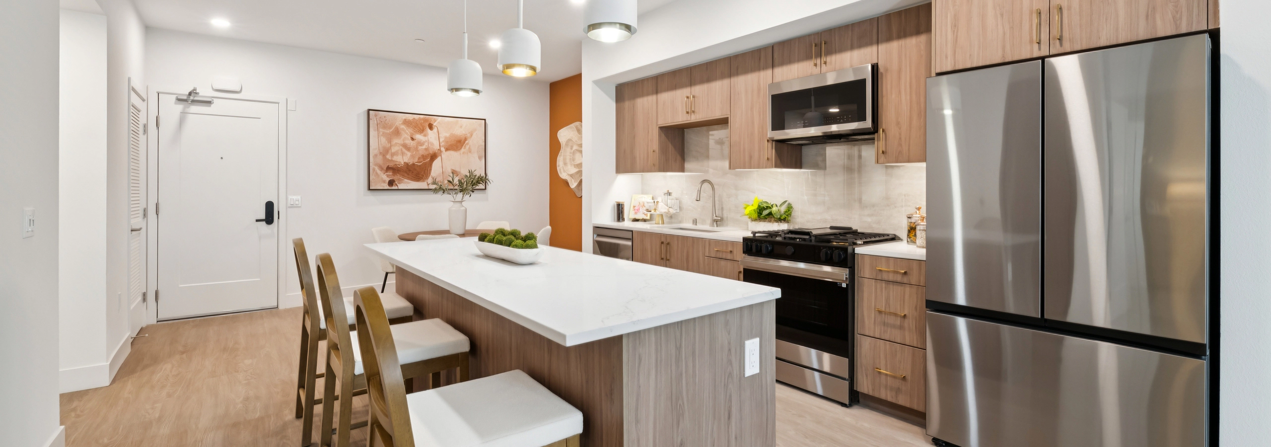 Open-concept kitchen and living area with wood cabinets, white island, bar stools, dining table, and pendant lights.