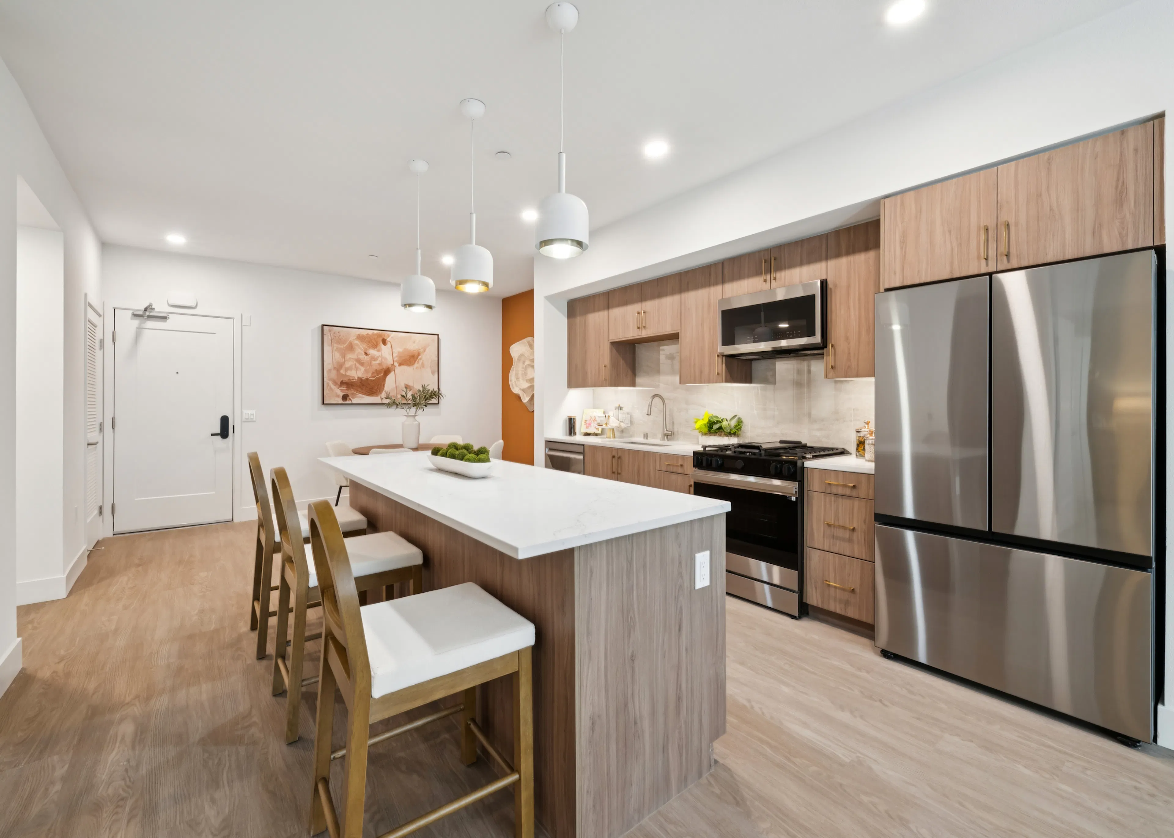 Open-concept kitchen and living area with wood cabinets, white island, bar stools, dining table, and pendant lights.