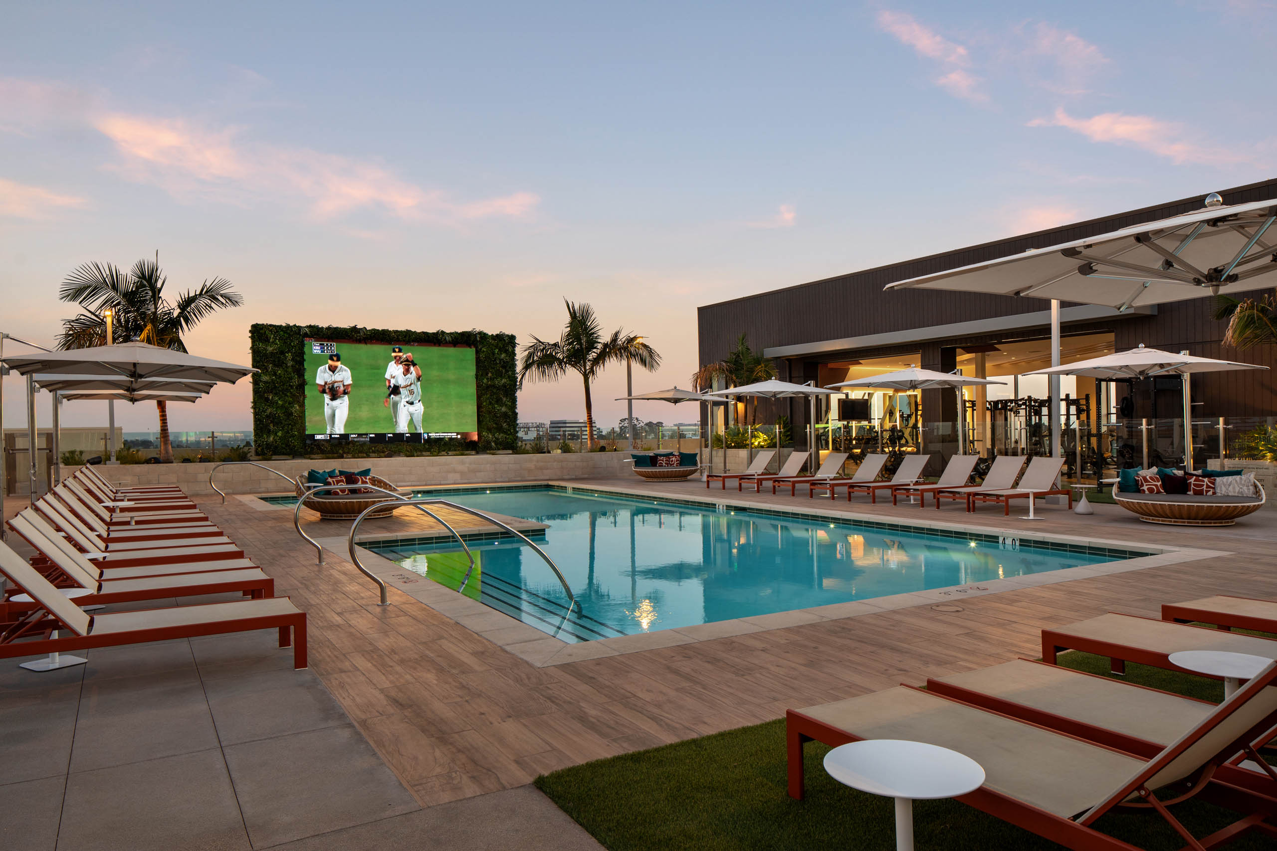 Resort-style outdoor pool with lounge chairs, umbrellas, large video screen, palm trees, fitness center, and deck at sunset.