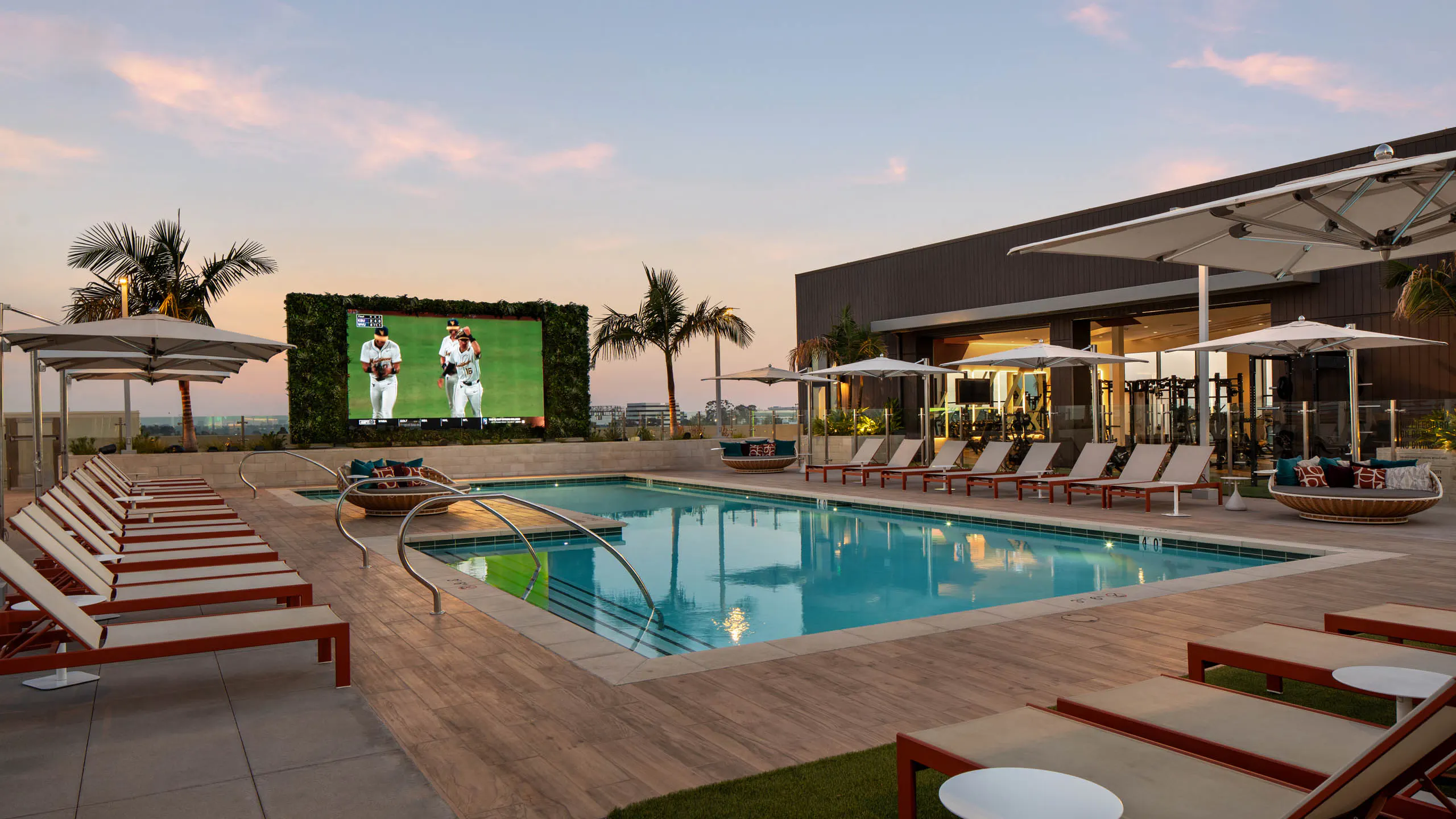 Resort-style outdoor pool with lounge chairs, umbrellas, large video screen, palm trees, fitness center, and deck at sunset.