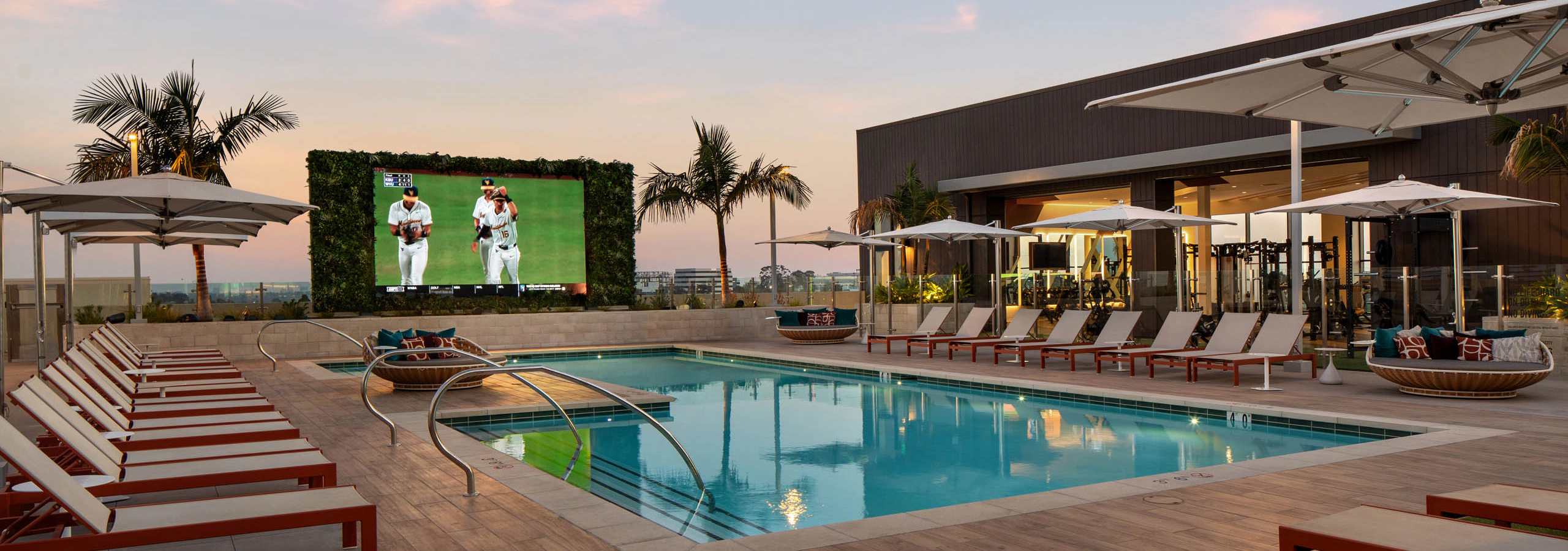 Resort-style outdoor pool with lounge chairs, umbrellas, large video screen, palm trees, fitness center, and deck at sunset.