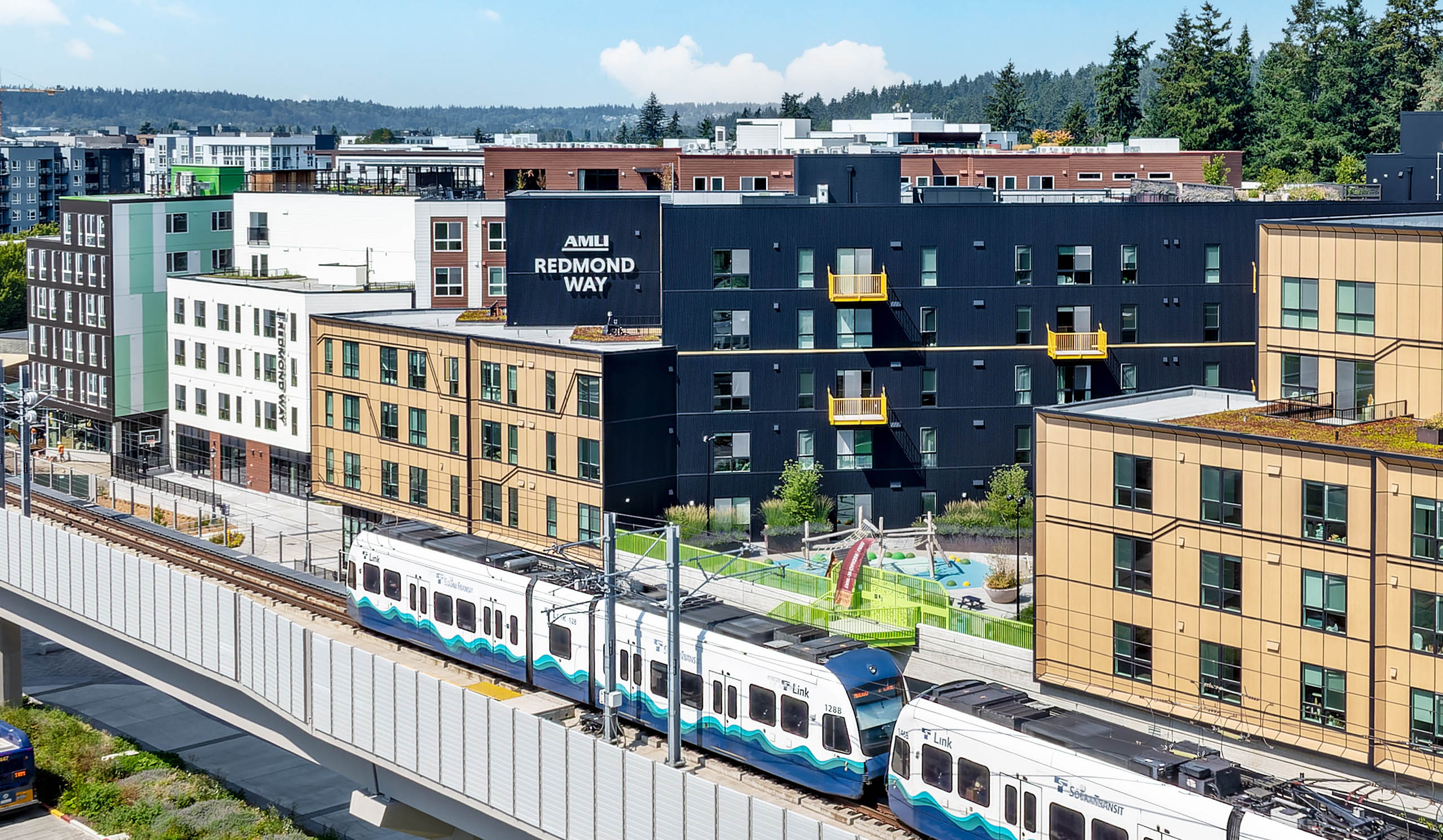 Aerial view of AMLI Redmond Way apartments with a Sound Transit Link light rail train passing alongside the community's outdoor play area and surrounding Redmond skyline.