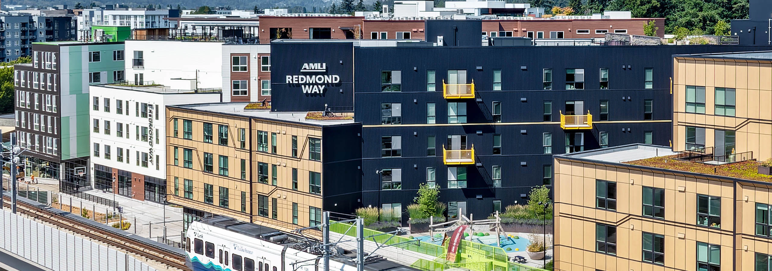 Aerial view of AMLI Redmond Way apartments with a Sound Transit Link light rail train passing alongside the community's outdoor play area and surrounding Redmond skyline.