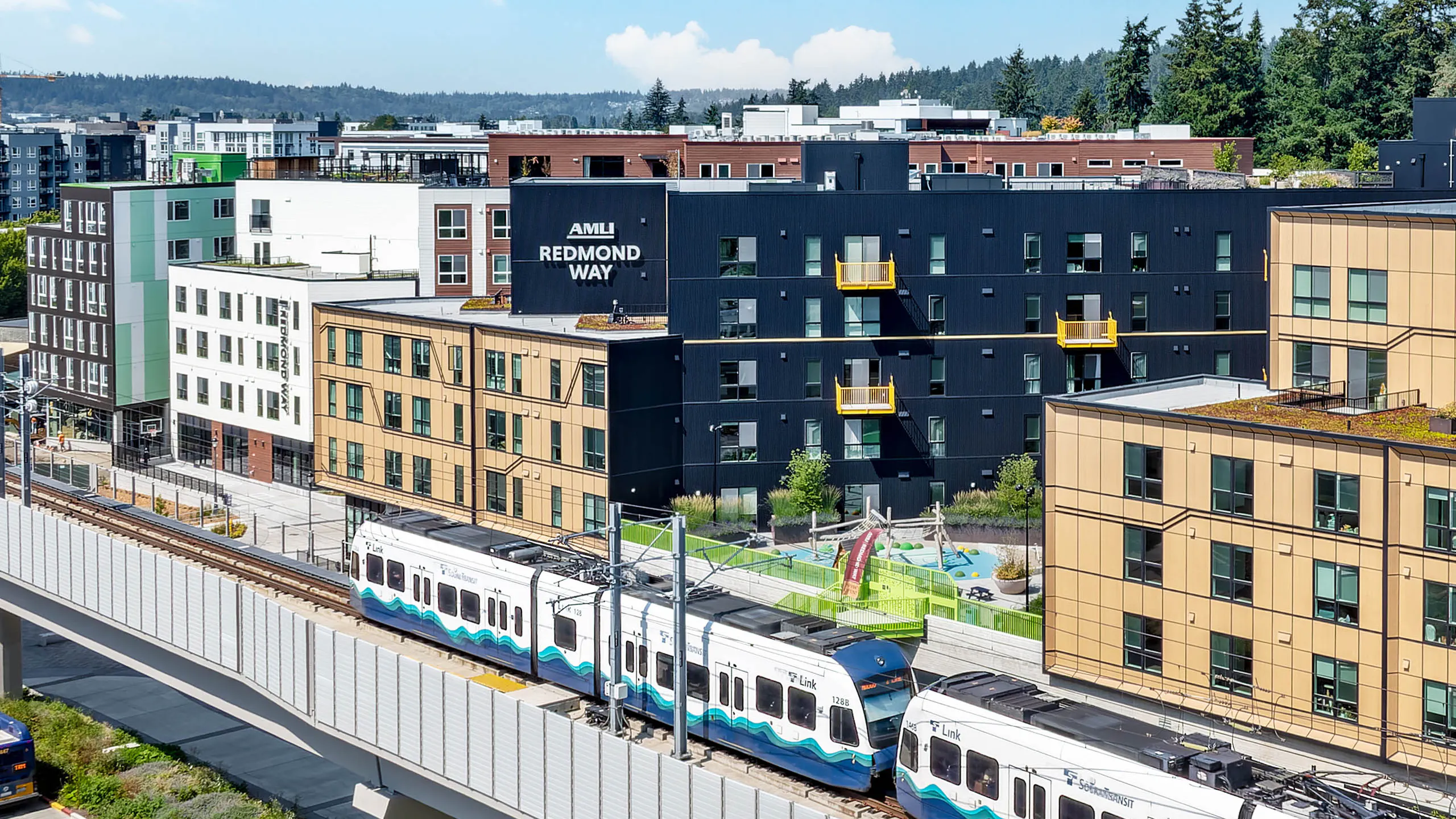 Aerial view of AMLI Redmond Way apartments with a Sound Transit Link light rail train passing alongside the community's outdoor play area and surrounding Redmond skyline.