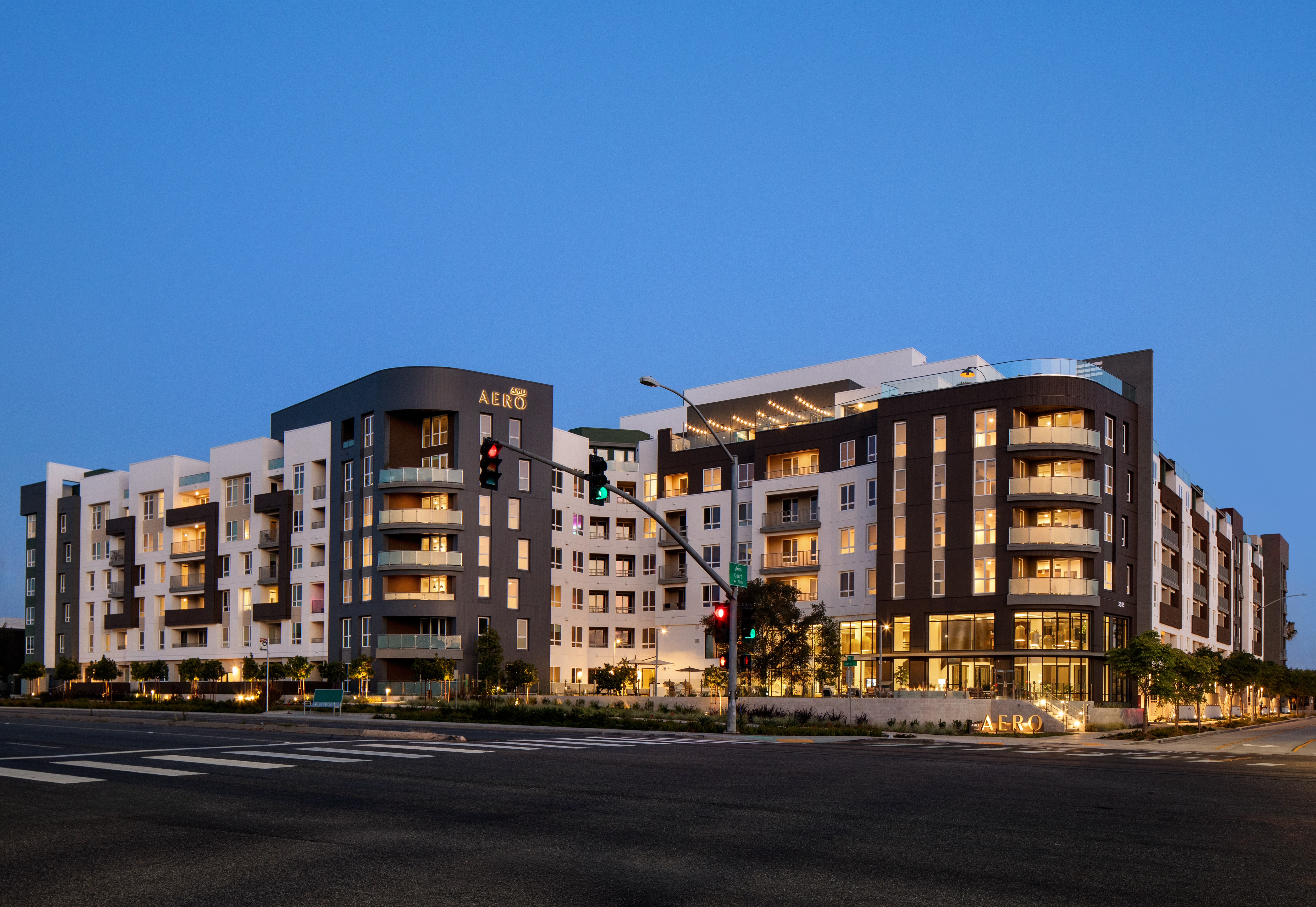 Exterior view of AMLI Aero at dusk with illuminated balconies, large windows, landscaped grounds, and a corner entrance.