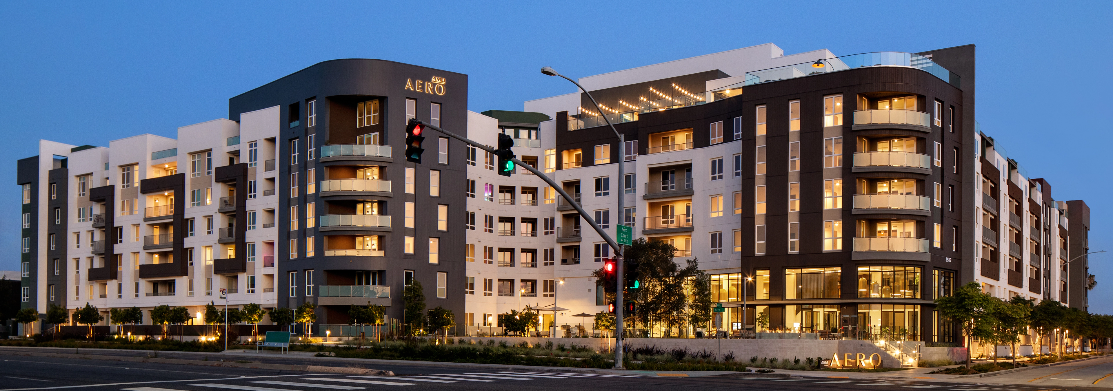 Exterior view of AMLI Aero at dusk with illuminated balconies, large windows, landscaped grounds, and a corner entrance.