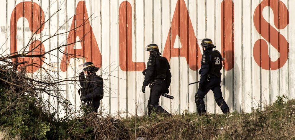 French riot police officer walk in front of a fence with the lettering 'Calais' near the A16 motorway near the site of the Eurotunnel in Coquelles, near Calais, northern France on January 21, 2016. - Approximately 300 migrants have tried to board trucks protected by French police, according to an estimate made by an AFP photographer present at the scene. Clashes already erupted briefly on the night of January 20 at the port bypass Calais between several hundred migrants and security forces, who fired tear gas to restore the situation, according to an AFP correspondent. These incidents occurred after the prefecture of Pas-de-Calais had set an ultimatum which expired early in the afternoon for the last migrants to leave a deforested 100 metre strip of the "Jungle" camp along the ring road for safety reasons. (Photo by PHILIPPE HUGUEN / AFP)