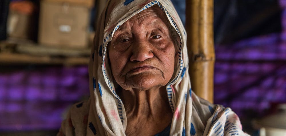  	Sokhina Khatun, a Rohingya woman around 90 years old, stands for a portrait in her shelter in Camp #1 East (Kutupalong Camp), Bangladesh, 19 February 2019.

“I’ve fled [from Myanmar] four times in my life," she told Amnesty International. "The fourth time was in 2017. I came here with only my walking stick [and] this thami (longyi)… There has never been peace for us. We’ve had to flee here frequently. We were under persecution for so long.”

“My number one problem [in the camp] is the latrine. The latrine is down at the bottom [of the hill], it’s very difficult for me to go down there. Sometimes I just go inside [my shelter in a pan]… I’ve been surviving like that.” 