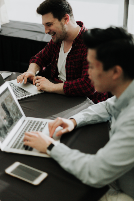 photo of two laughing men sitting at a table working on their laptops 