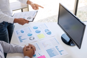Primer plano de analistas de negocios revisando gráficos financieros y documentos sobre un escritorio, con un monitor apagado al fondo.