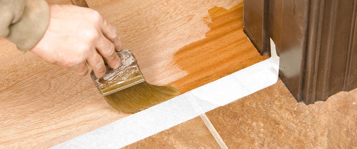 Using a paintbrush to apply a coat of finish to chemically buffed hardwood floors around baseboards and the edges of the room.