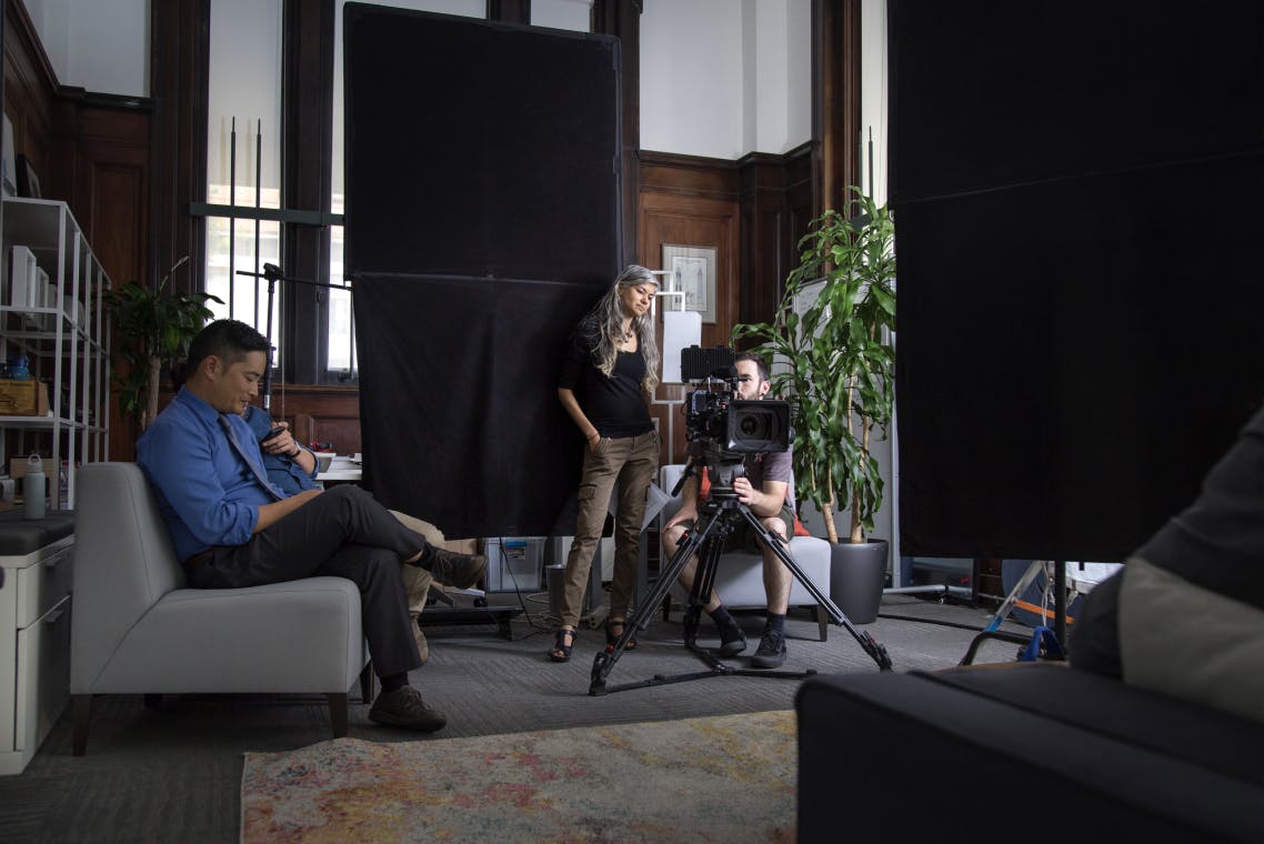 Woman with long gray hair stands next to a seated man with brown hair, they both are watching a viewfinder on a video shoot. On the left are two actors sitting on a couch and looking at their phones.