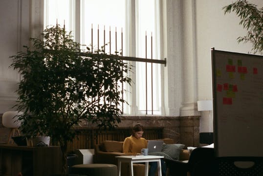 Woman in yellow sweater sits alone on a couch working on her laptop