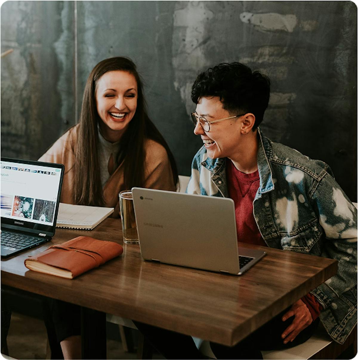 Duas mulheres sorrindo em frente ao computador. Elas estão utilizando roupas normais, uma possuí um cabelo longo a sua colega cabelo curto e óculos.