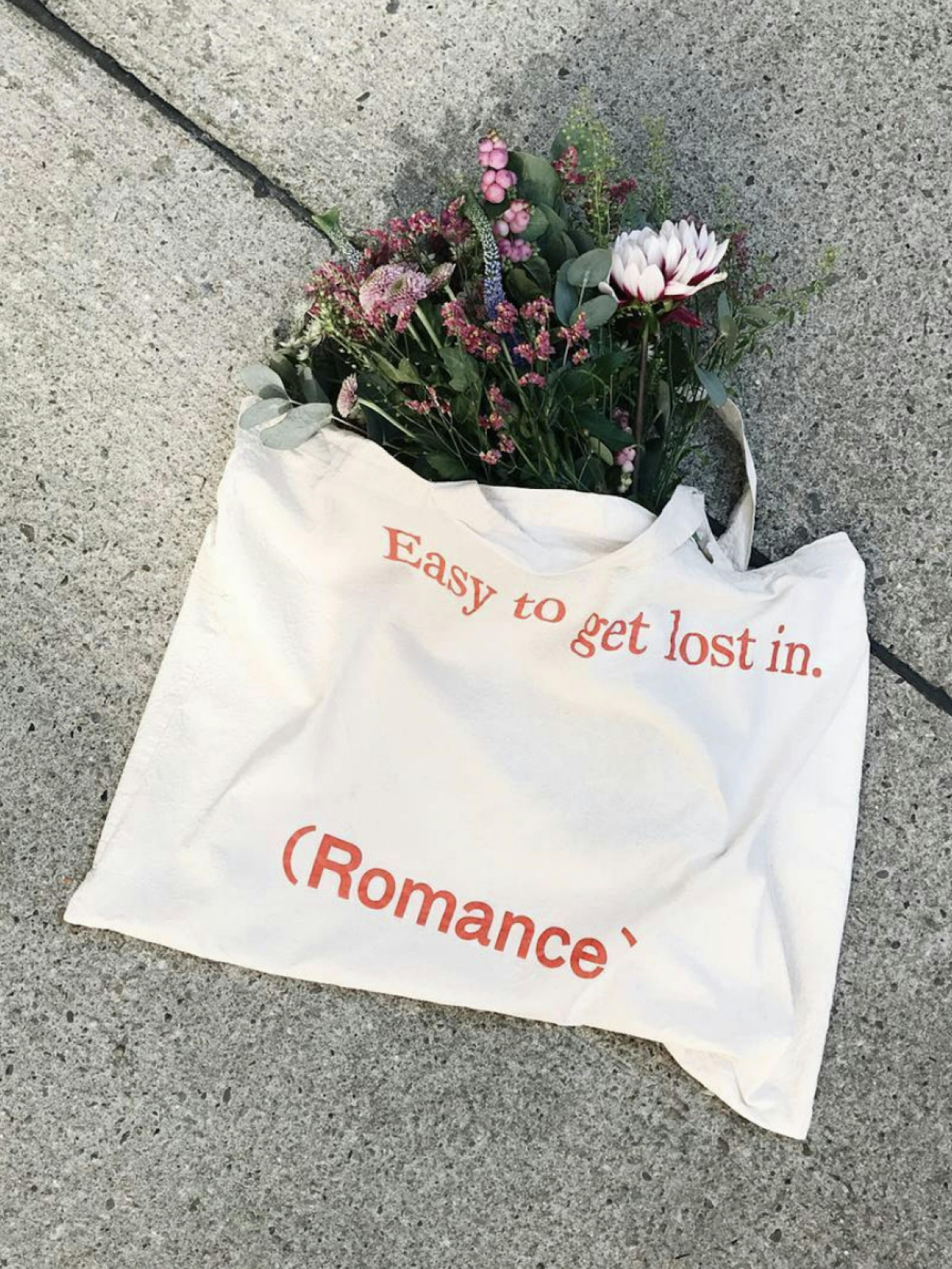 mint & berry by Zalando – Brand Campaign "The world needs more romance": Photo showing a white tote bag with orange text "Easy to get lost in. (Romance)" filled with a bouquet of pink and white flowers, lying on a concrete surface x HY.AM Studios