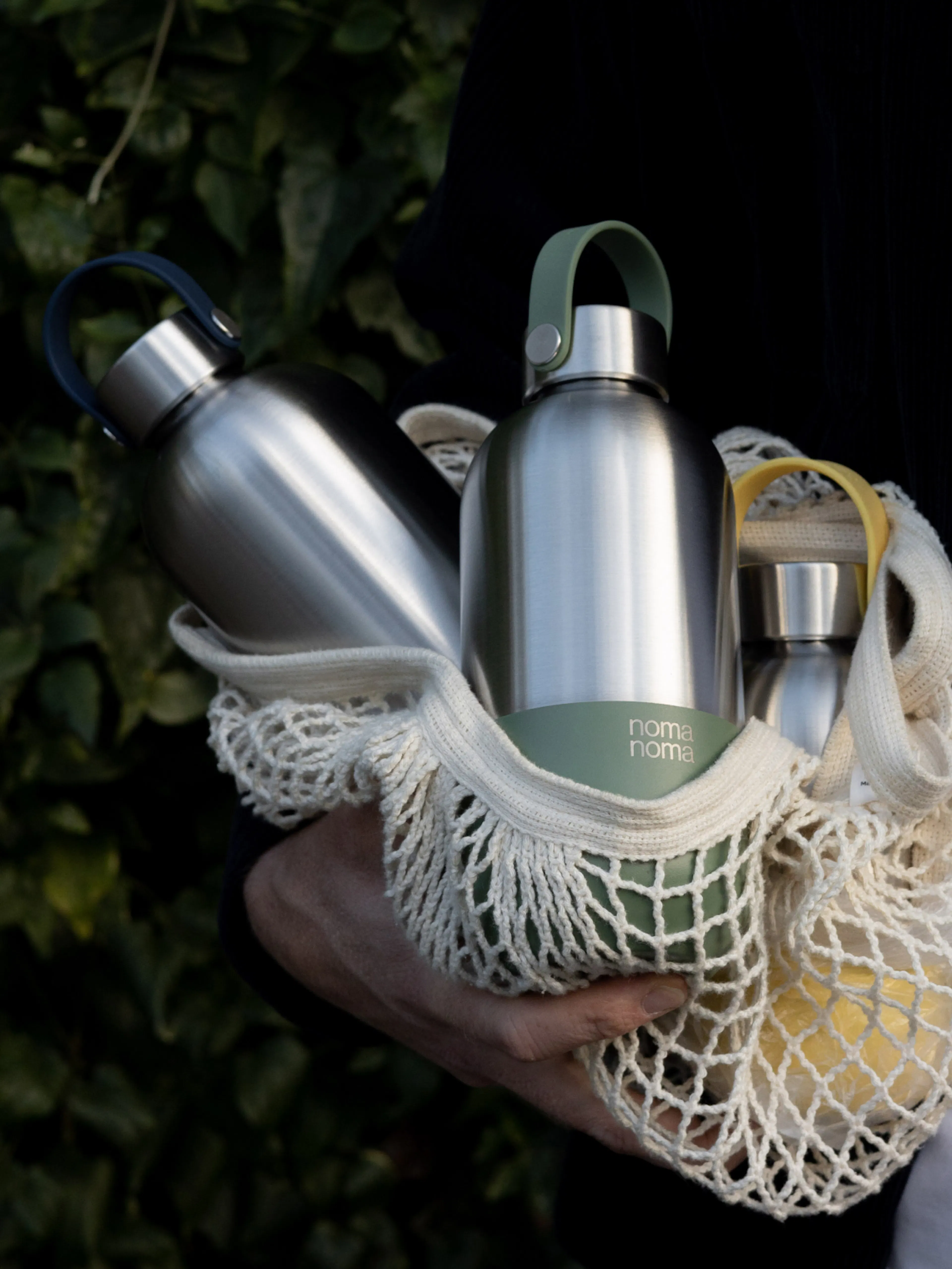 noma noma – Brand Design, E-Commerce, Experience Design "A conscious companion becomes a personal style icon": Photo showing a person holding a net bag with three stainless steel water bottles, two with colored loops x HY.AM Studios