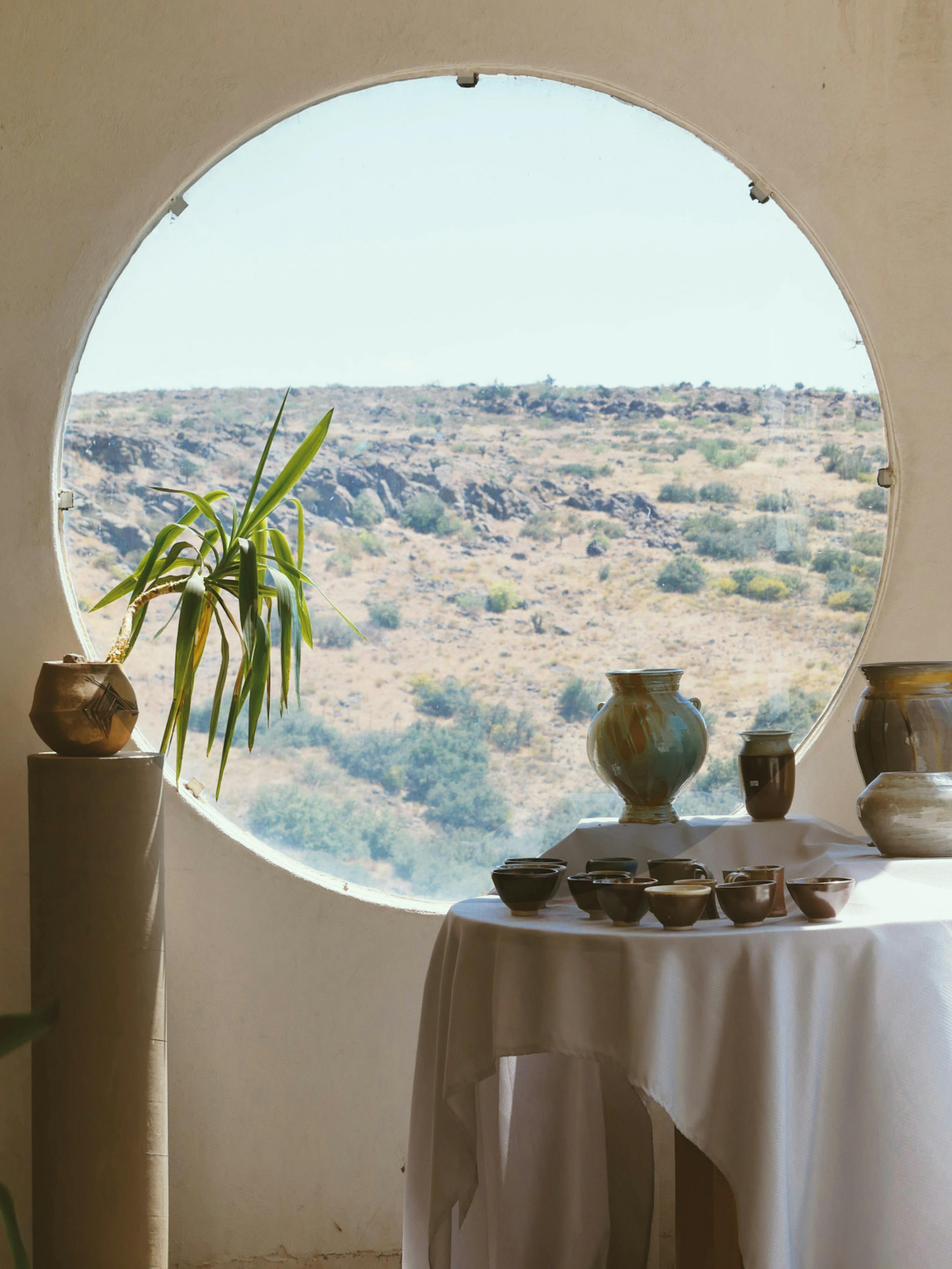 Creme Guides – Brand Design "Discovering Europe with style": Photo showing a collection of ceramic vases and bowls displayed on a table with a white tablecloth, framed by a circular window overlooking a dry, hilly landscape x HY.AM Studios