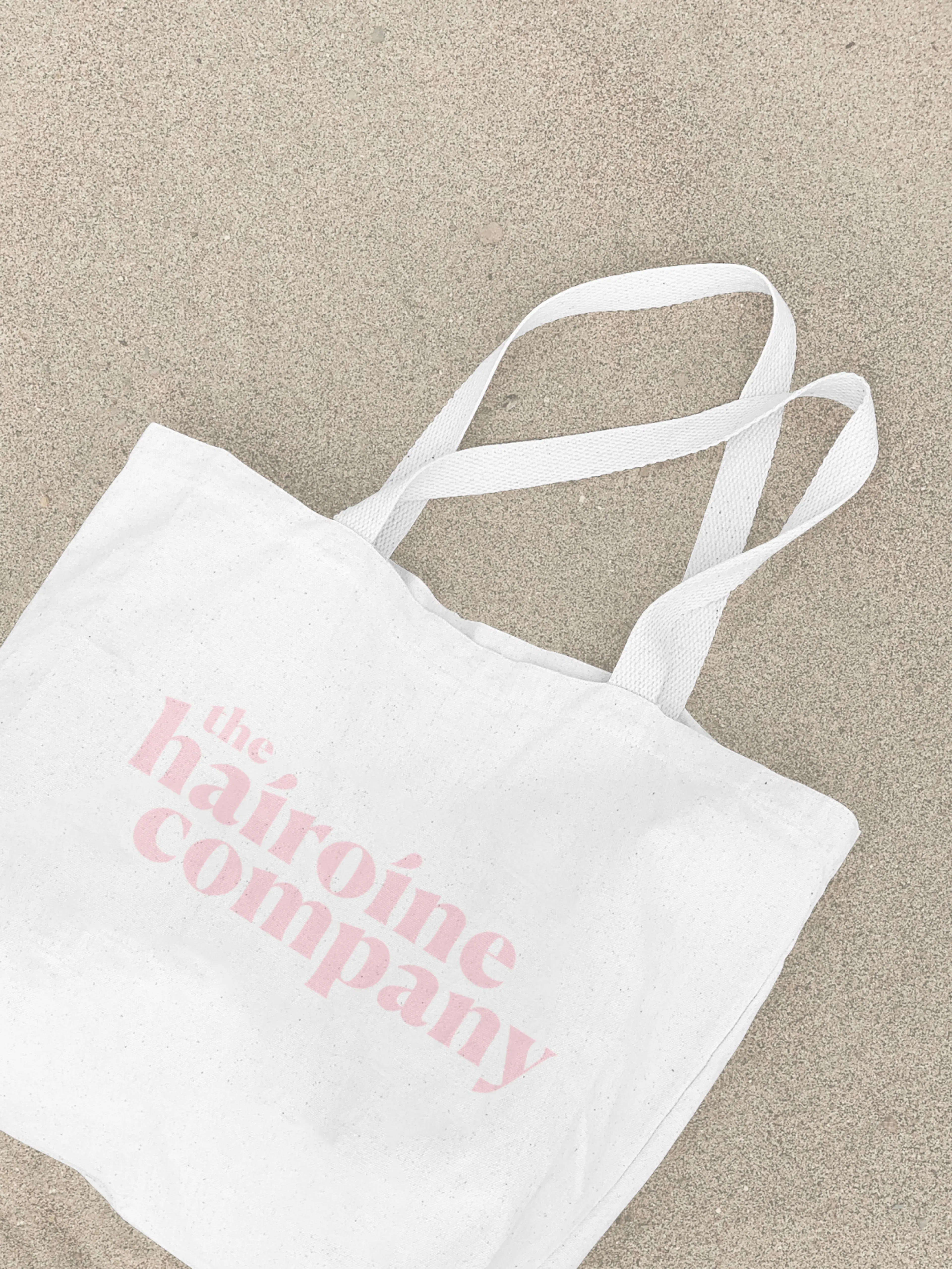 The Hairoine Company – Branding "A hair care brand for confident women": Mockup showing a white tote bag with pink logo on sand by HY.AM