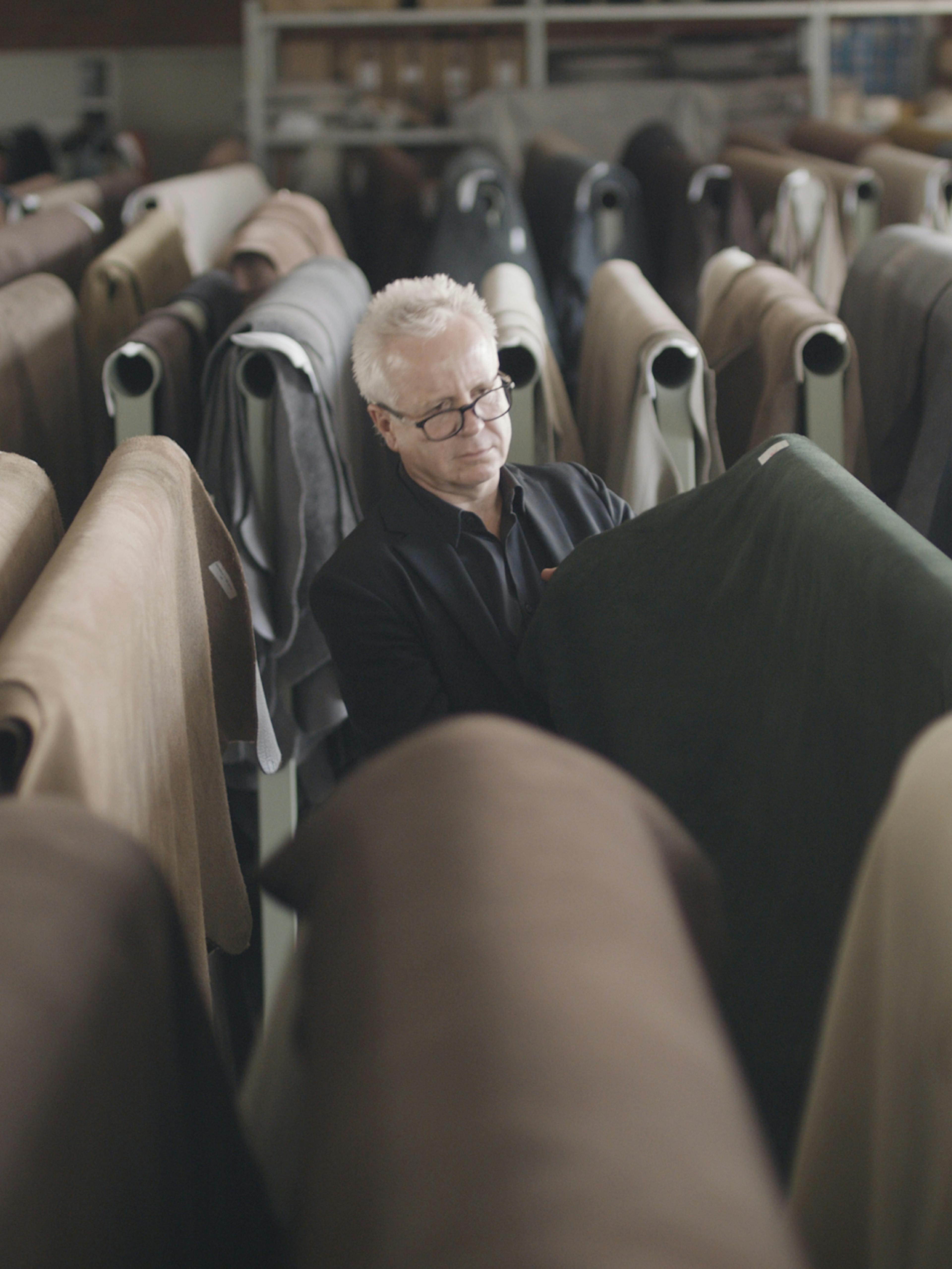Walter Knoll – Brand Design, E-Commerce, Experience Design "The essence of excellence": Photo showing a man in glasses examining a roll of dark green fabric in a warehouse filled with rolls of material x HY.AM Studios