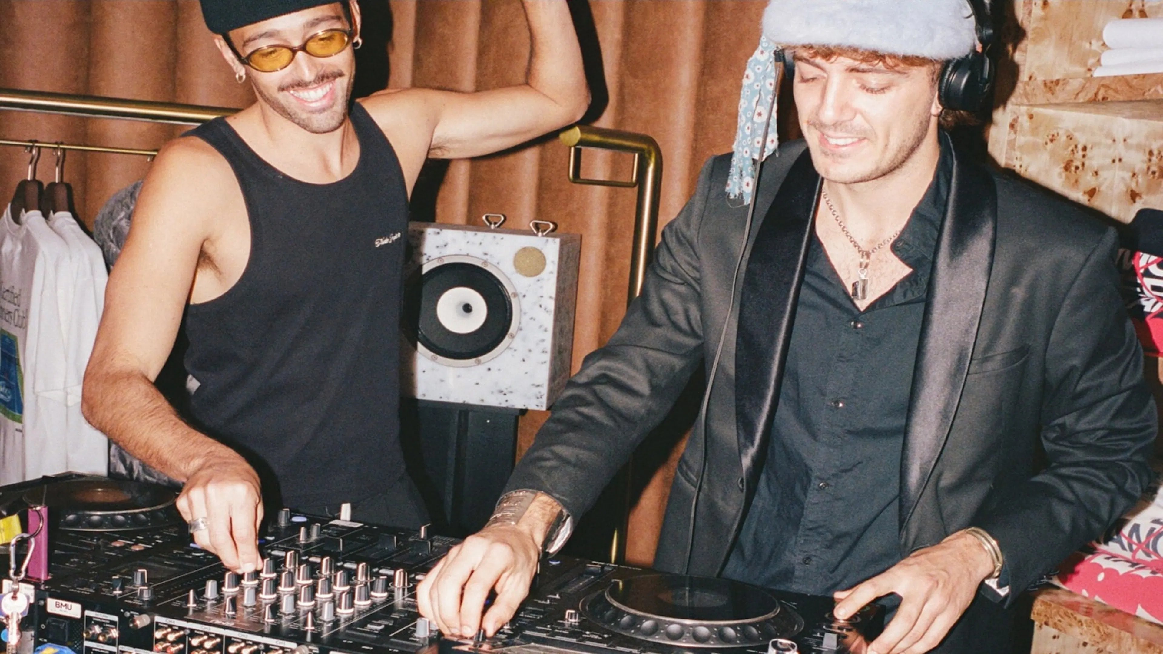 Nude Project - Communications : Photo showing Two DJs mixing music at a lively in-store event, surrounded by clothing racks and speakers.